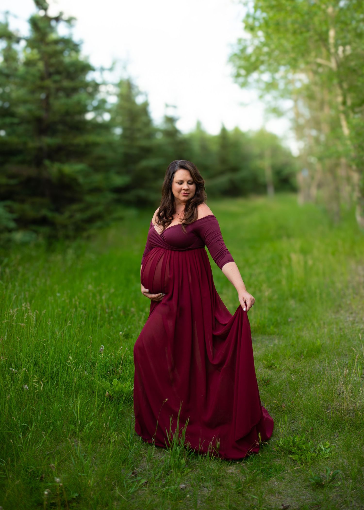 Pregnant woman in burgundy off-shoulder gown poses gracefully in lush green meadow surrounded by trees.