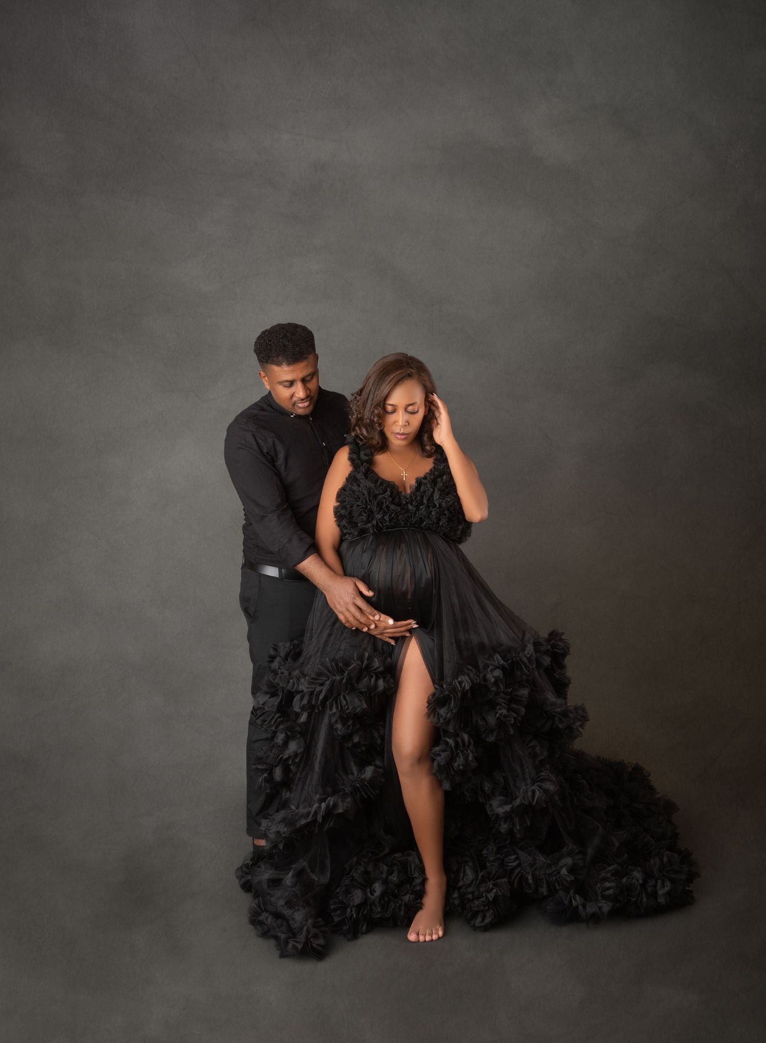 Couple in elegant black maternity photoshoot, woman in ruffled black gown with man in black outfit on gray backdrop.