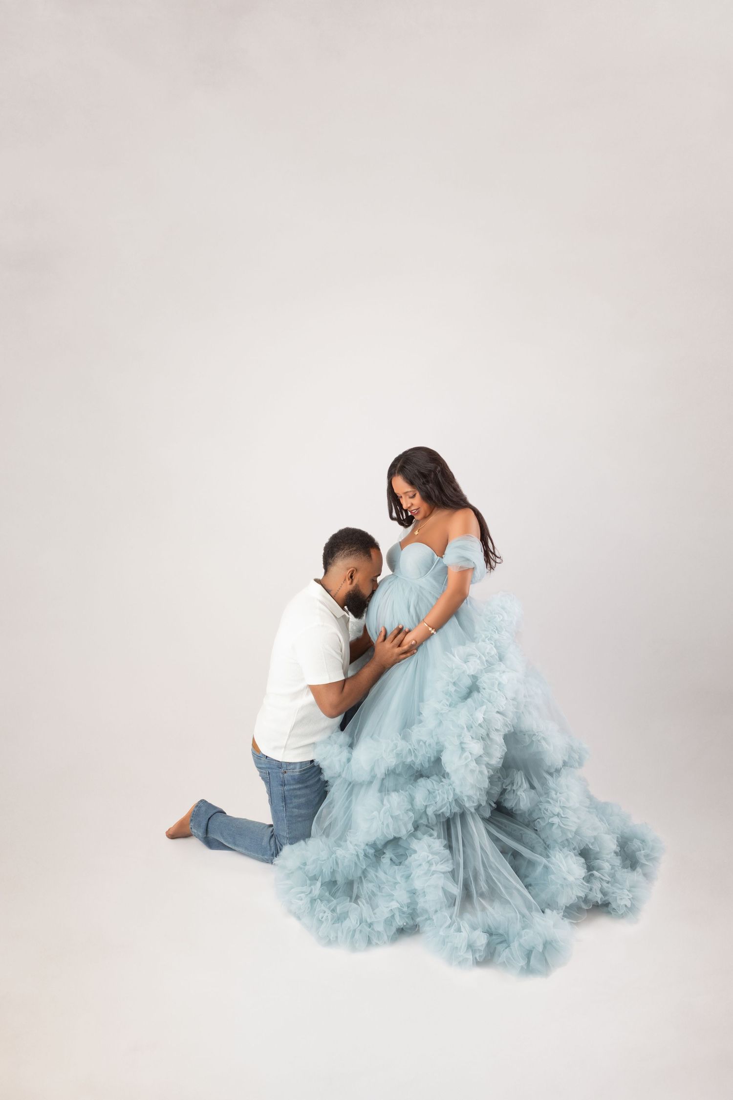 Man kneeling before pregnant woman in flowing blue tulle gown during maternity photoshoot on white background.