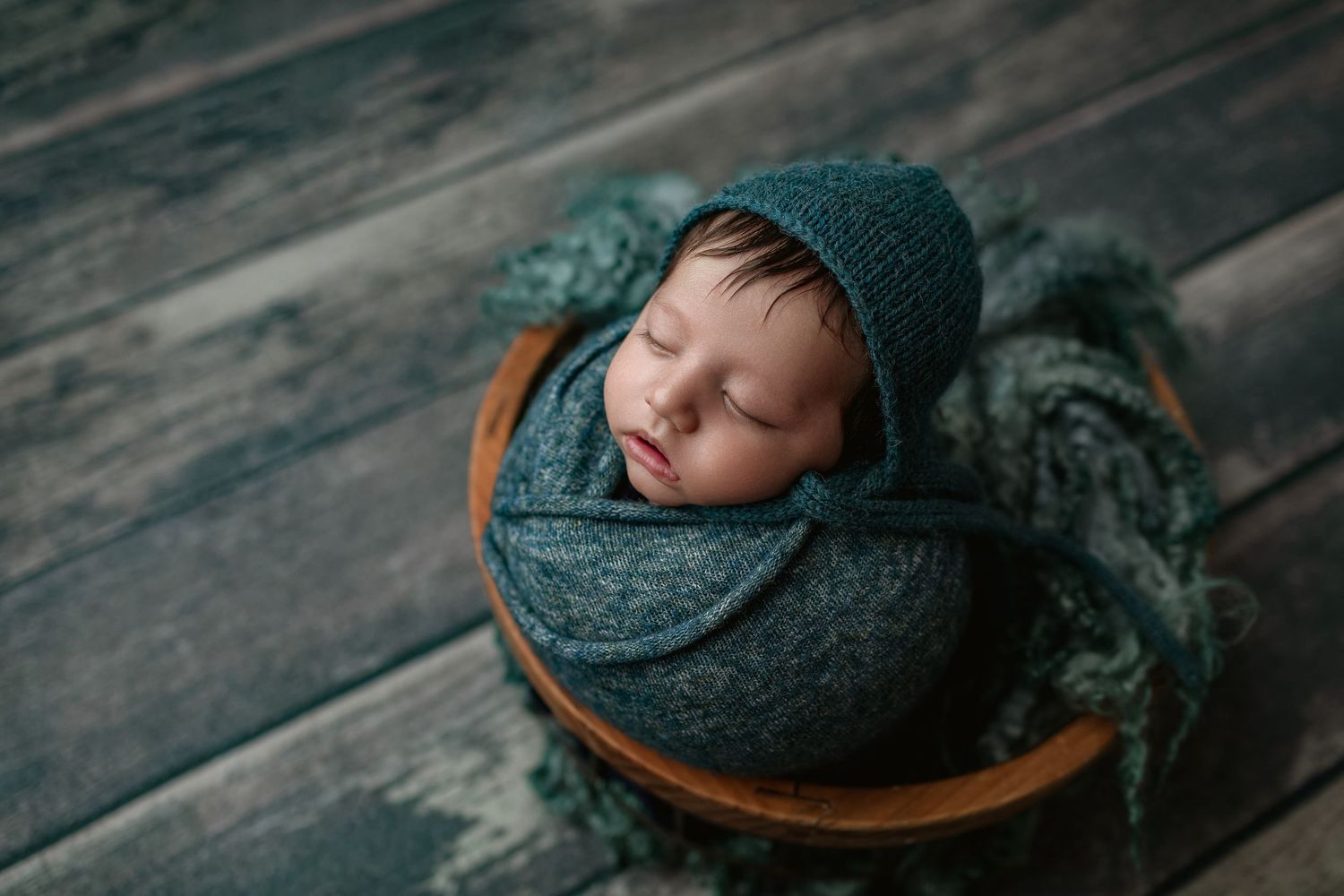 Sleeping newborn baby wrapped in teal knit blanket and bonnet, nestled in wooden bowl on rustic gray wood floor.