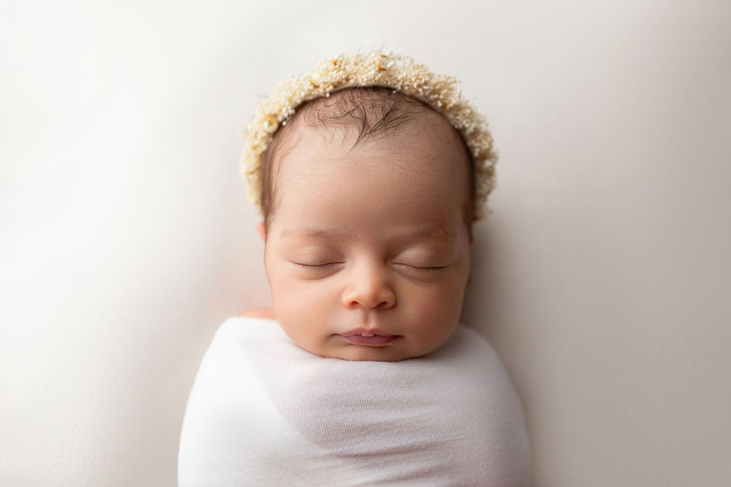 Peaceful newborn baby sleeping wrapped in white swaddle wearing a delicate floral headband on white background.