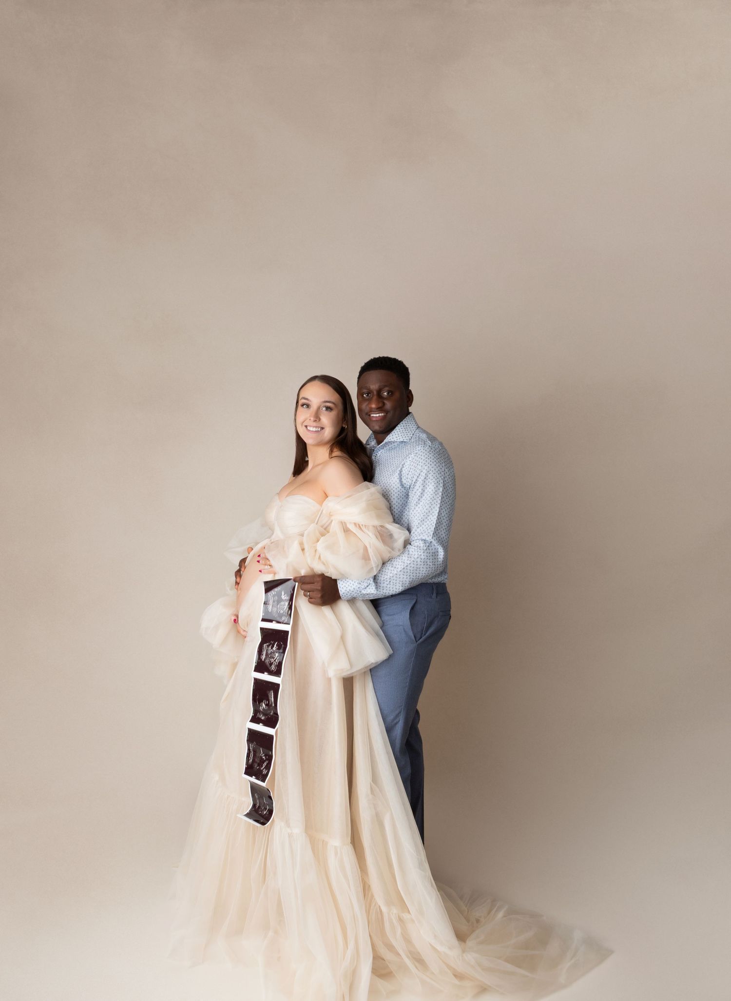Pregnant couple posing in studio, woman in flowing cream tulle gown holding ultrasound photos, man in blue suit embracing her.