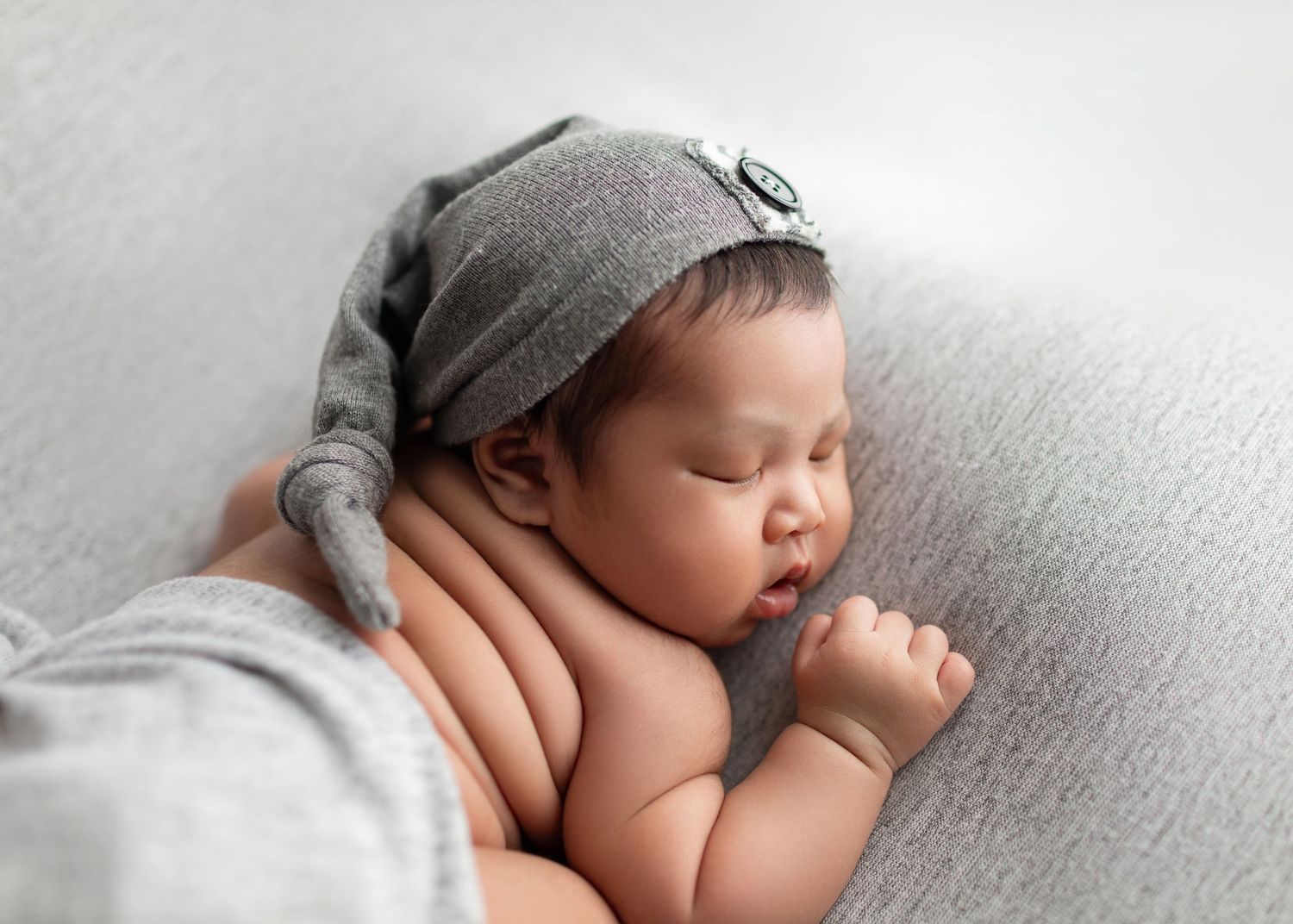 Sleeping newborn baby wearing a gray knotted hat nestled in a soft gray blanket.