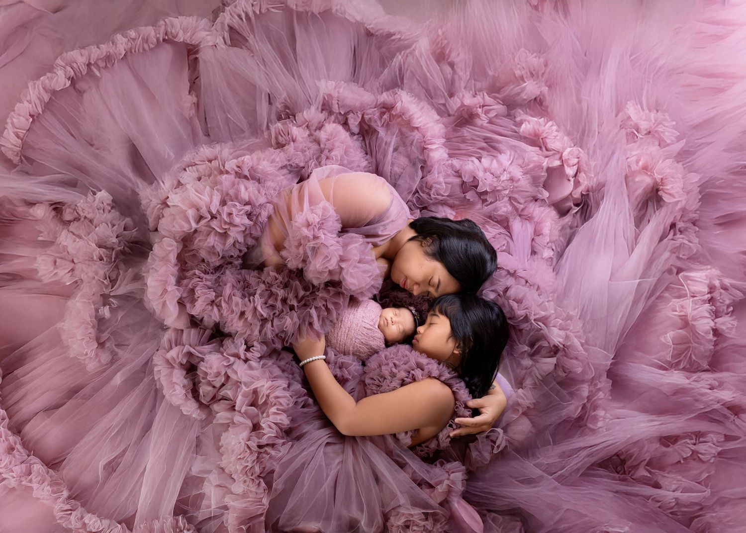 Aerial view of mom and daughter in purple tulle gowns cuddling newborn on ruffled fabric.