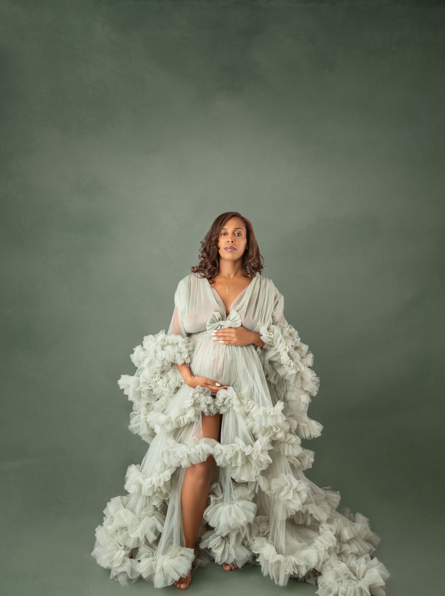 Pregnant woman in white ruffled maternity gown posing gracefully against a soft green studio background.