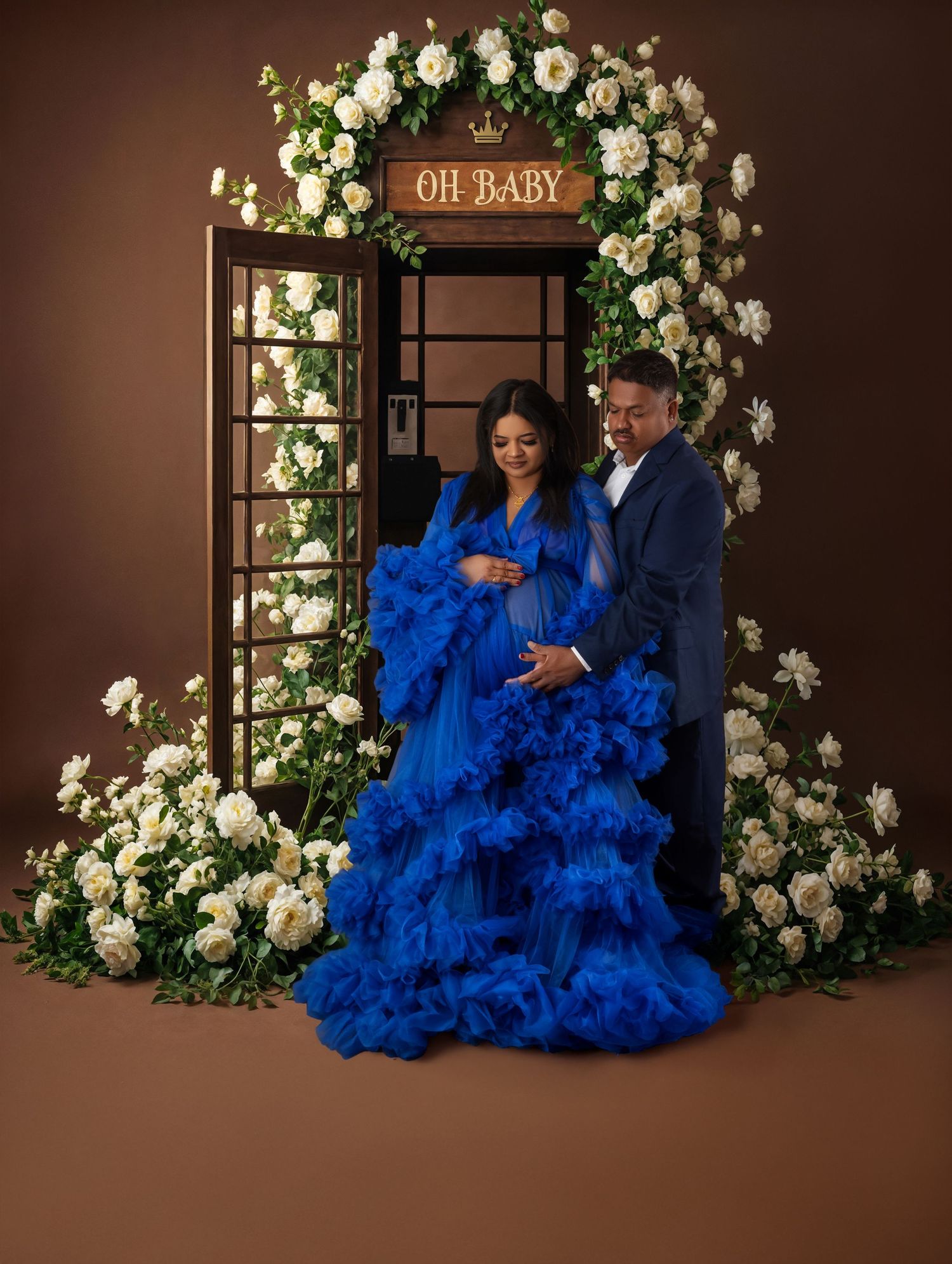 Couple in royal blue maternity gown poses before floral arch with OH BABY sign for elegant maternity photo.