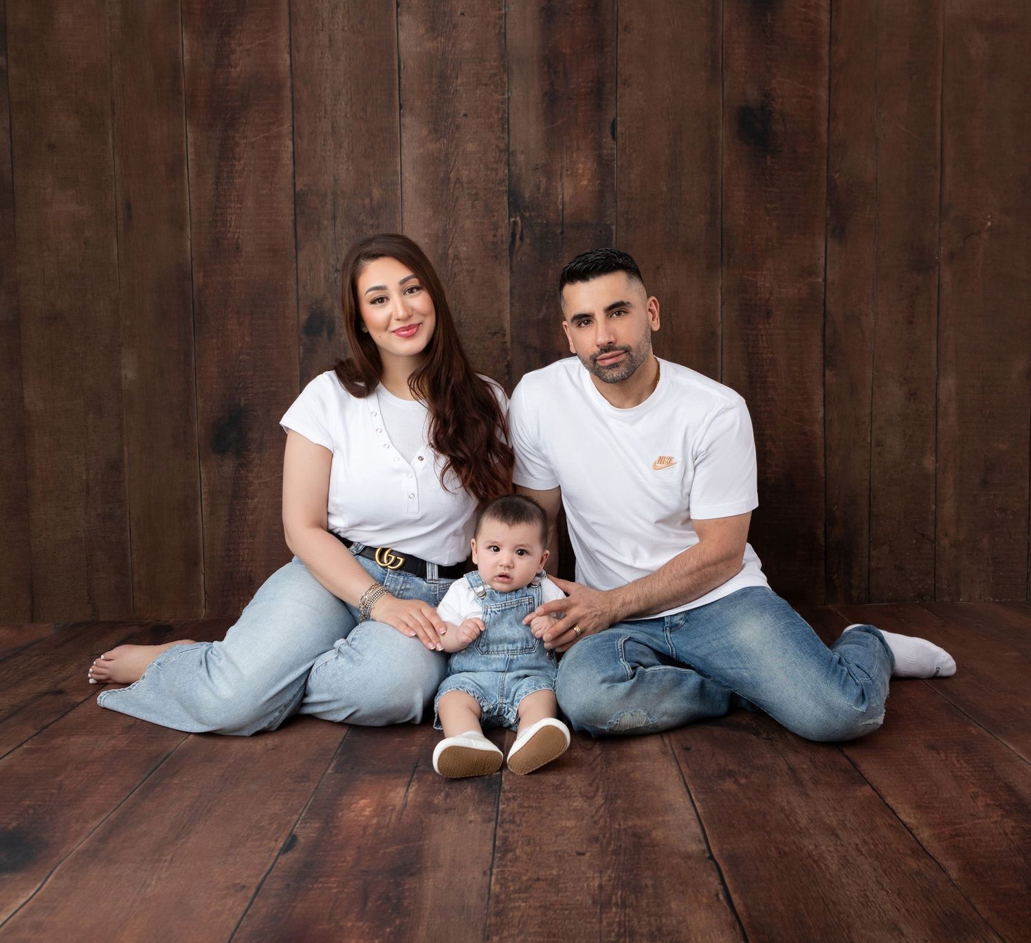 Young family with baby sitting on wooden floor against rustic wood panel backdrop in matching white tees and jeans.
