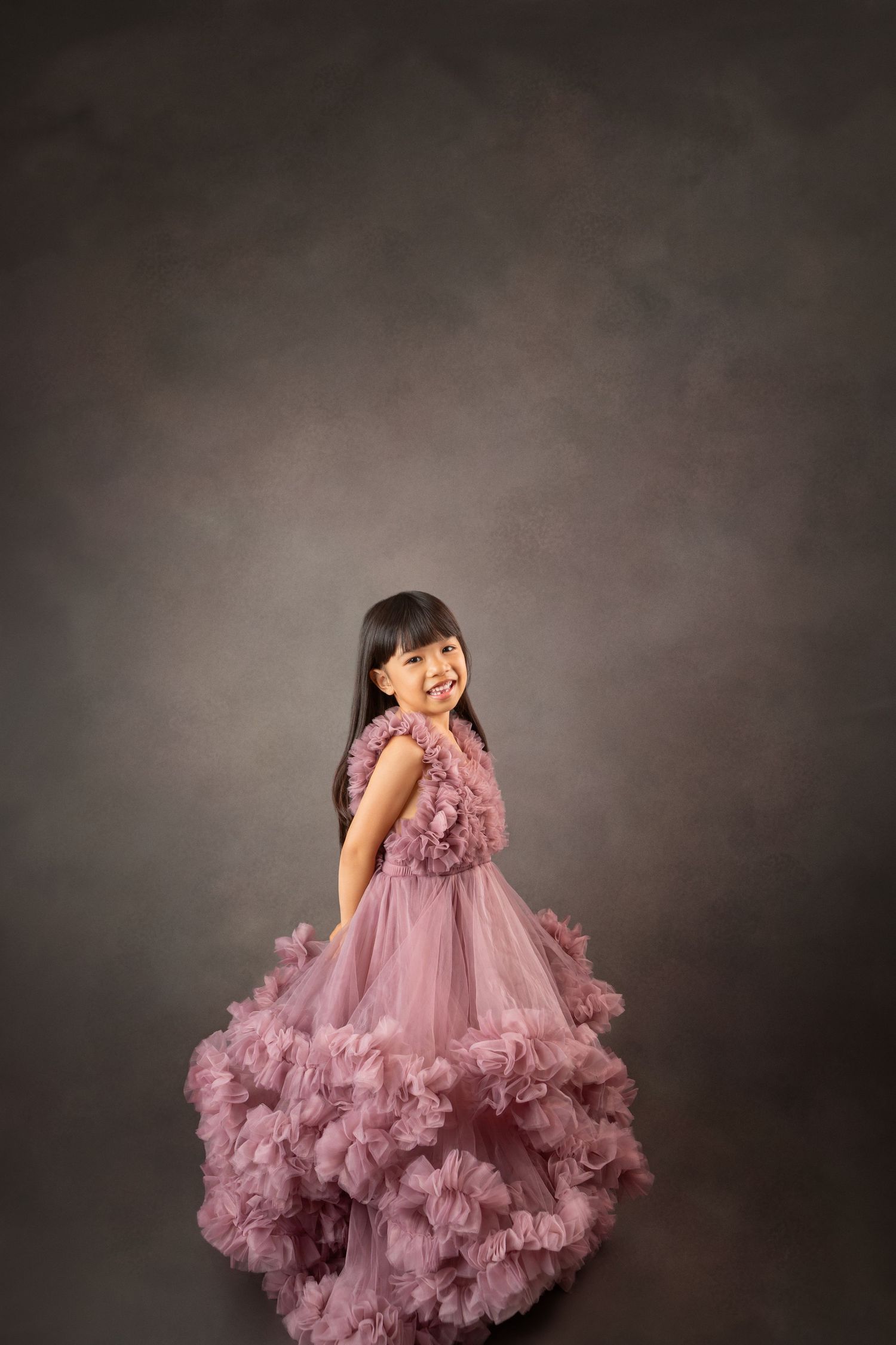A young girl twirls in a flowing pink ruffle gown, smiling over her shoulder against a gray studio backdrop.