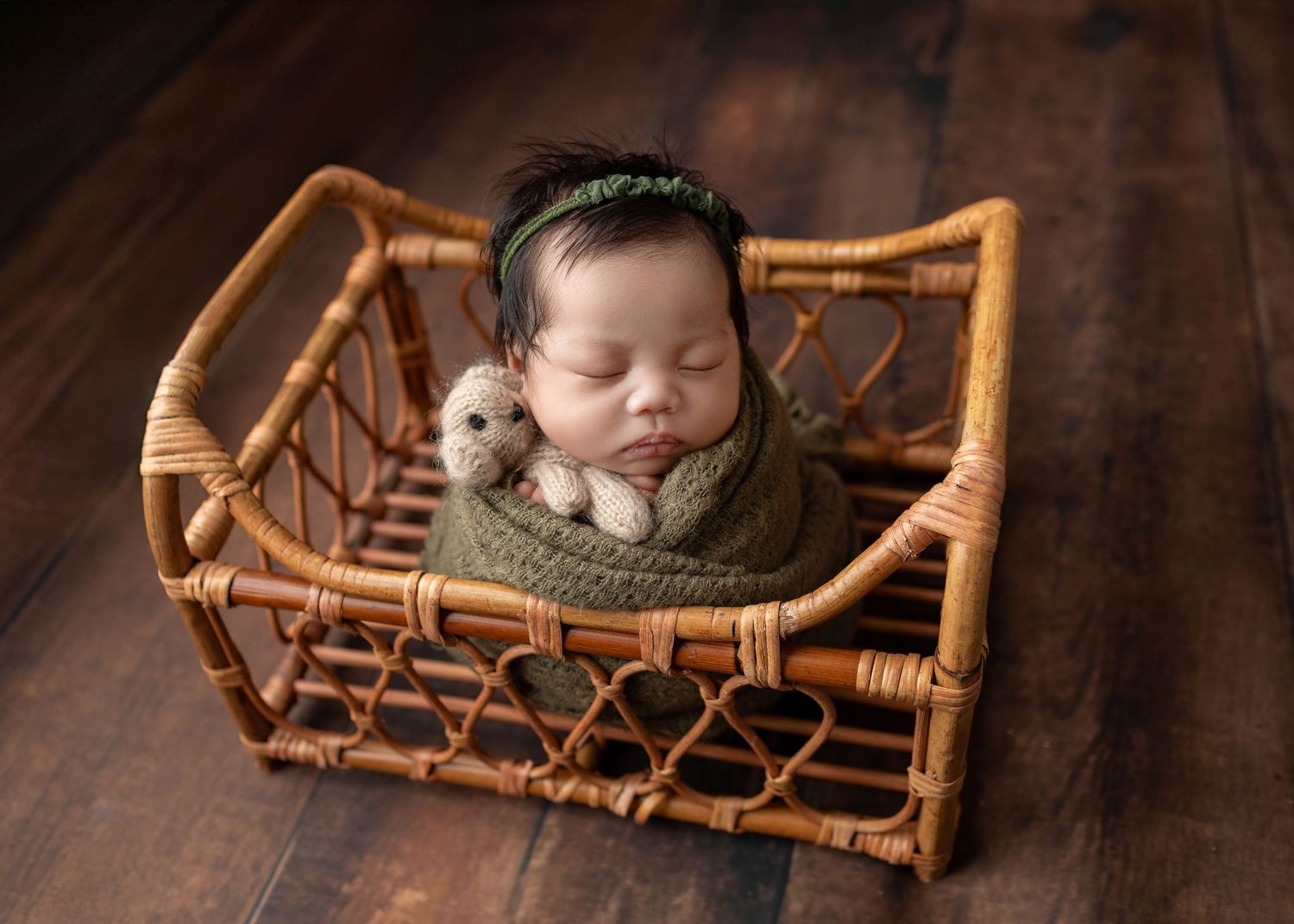 Newborn baby sleeping in a wicker basket wrapped in green knit, holding a tiny teddy bear, on a dark wood floor.