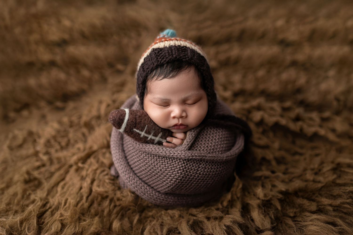 Sleeping newborn baby wrapped in brown knit swaddle with football prop and striped bonnet on fur backdrop.
