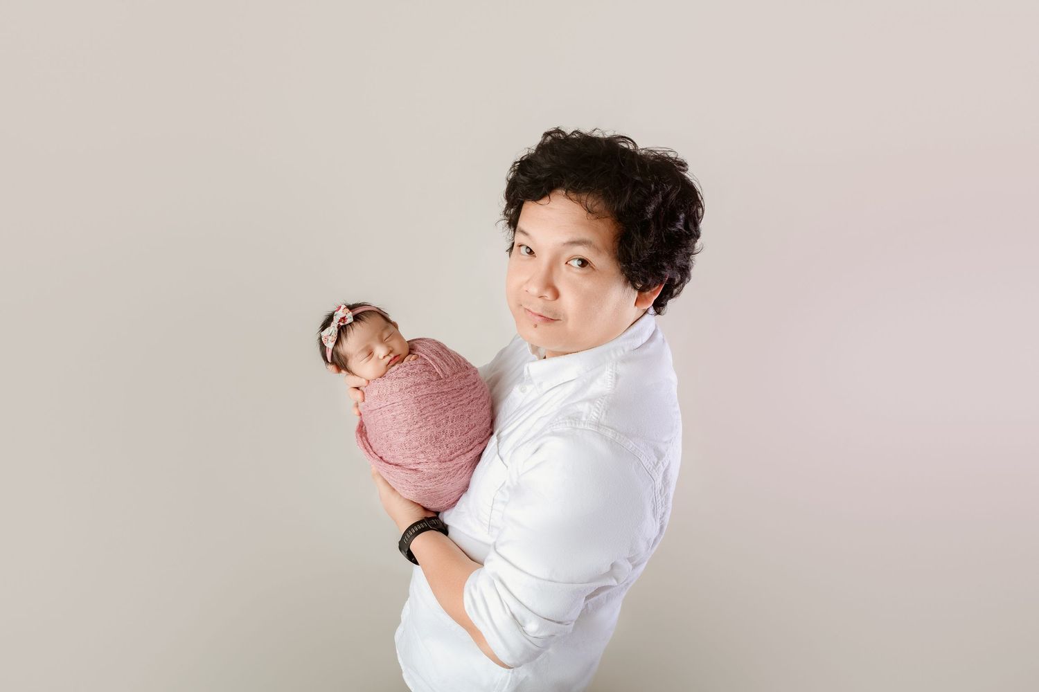 Parent in white shirt holding pink-swaddled newborn baby with headband over shoulder in studio portrait.