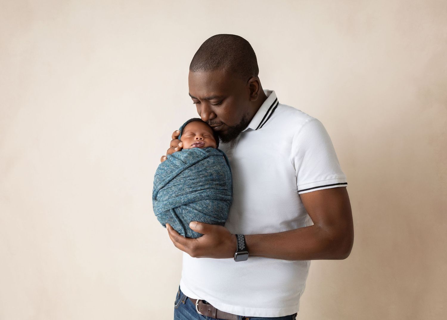 Loving father in white polo shirt tenderly kissing swaddled newborn baby against neutral beige background.