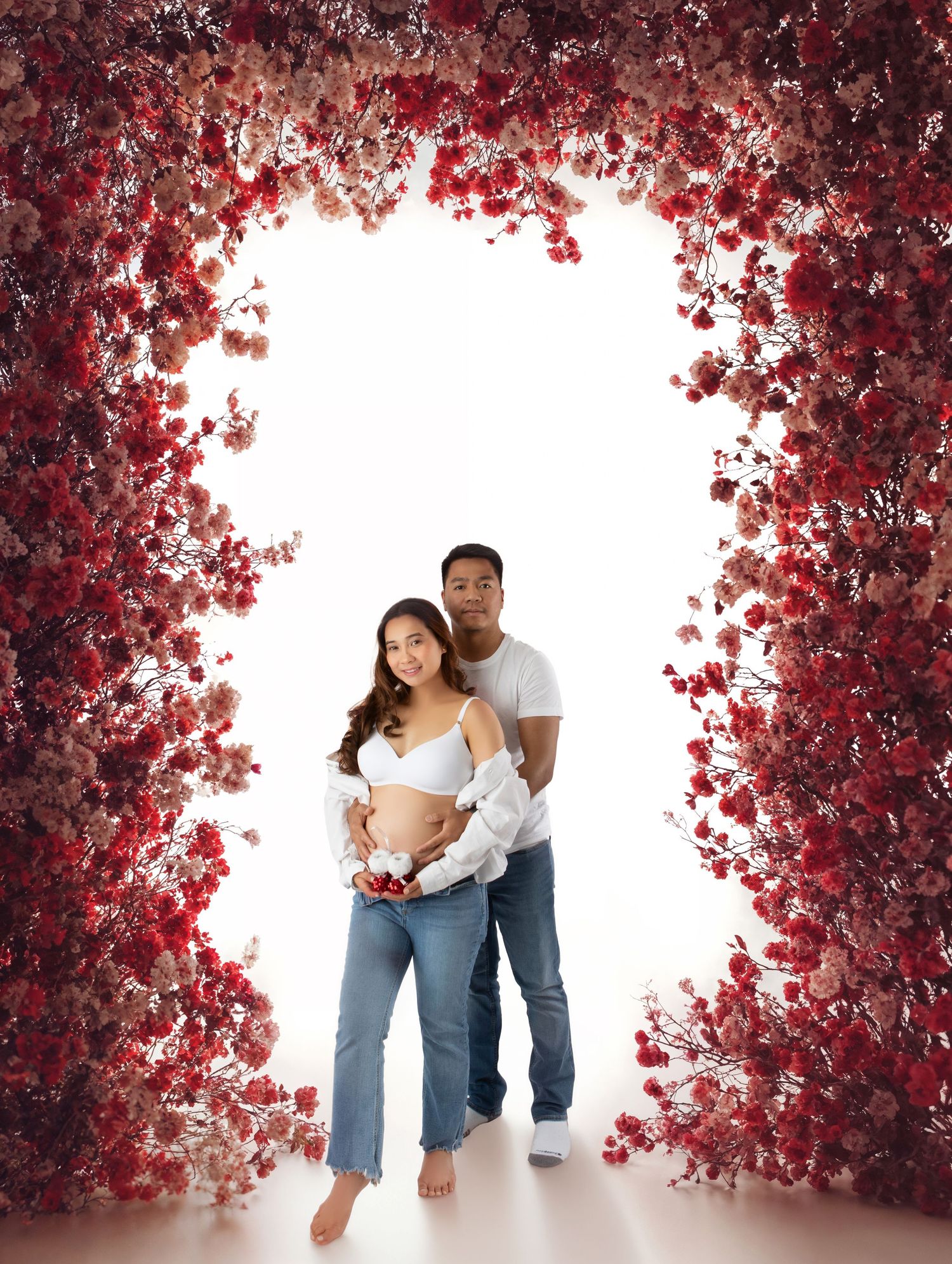 Pregnant couple stands under stunning red floral arch, woman in white bra top holding baby shoes.