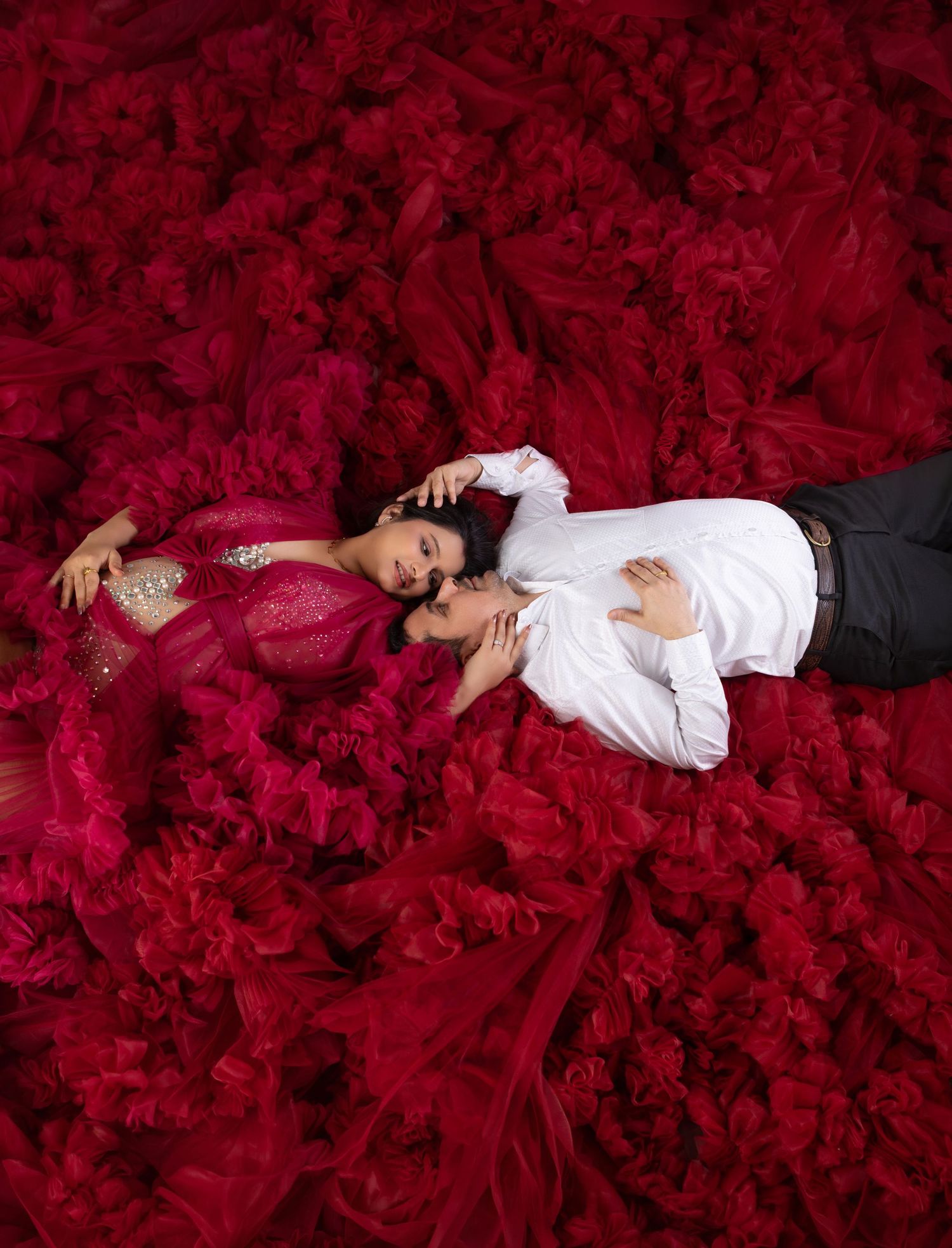 Couple lying together surrounded by a sea of dramatic red ruffled fabric in an artistic maternity portrait.