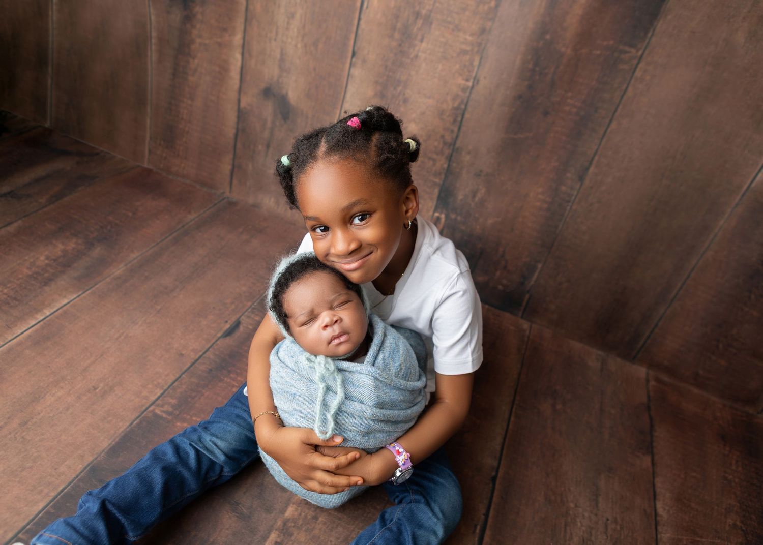 Young girl sitting against a wood wall lovingly holding her swaddled newborn sibling.