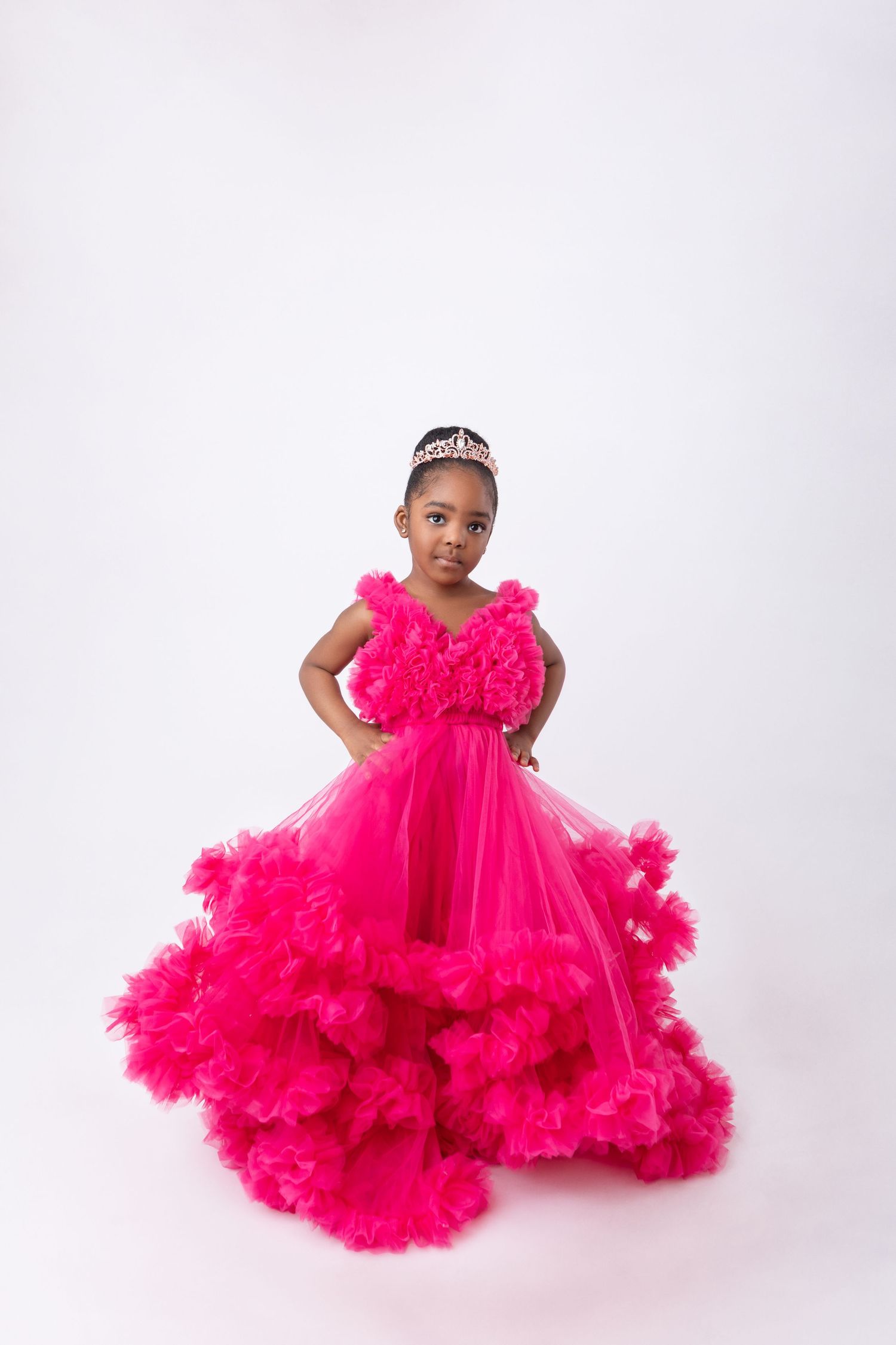 Young Black girl in a stunning hot pink ruffled tulle ball gown with tiara, posing confidently on white background.