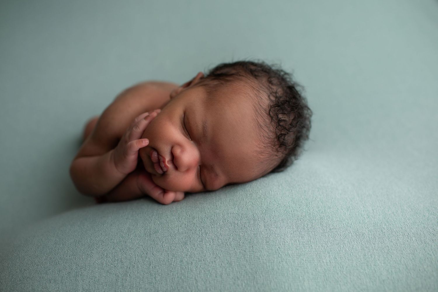 Newborn baby sleeping peacefully on a soft teal blanket, curled up with tiny hands near face.