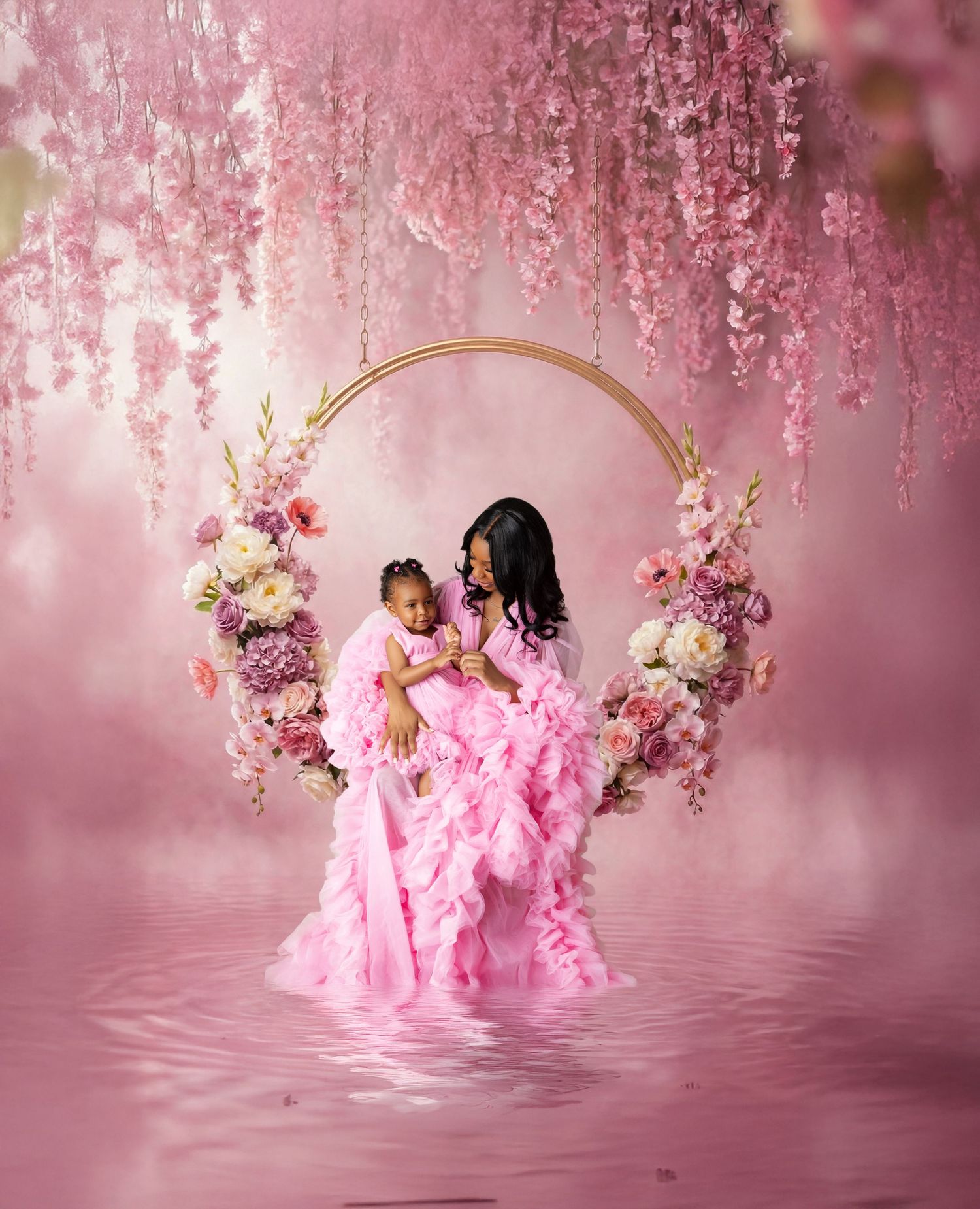 Mother and daughter in matching pink ruffle gowns sit on a floral arch swing surrounded by pink blooms.