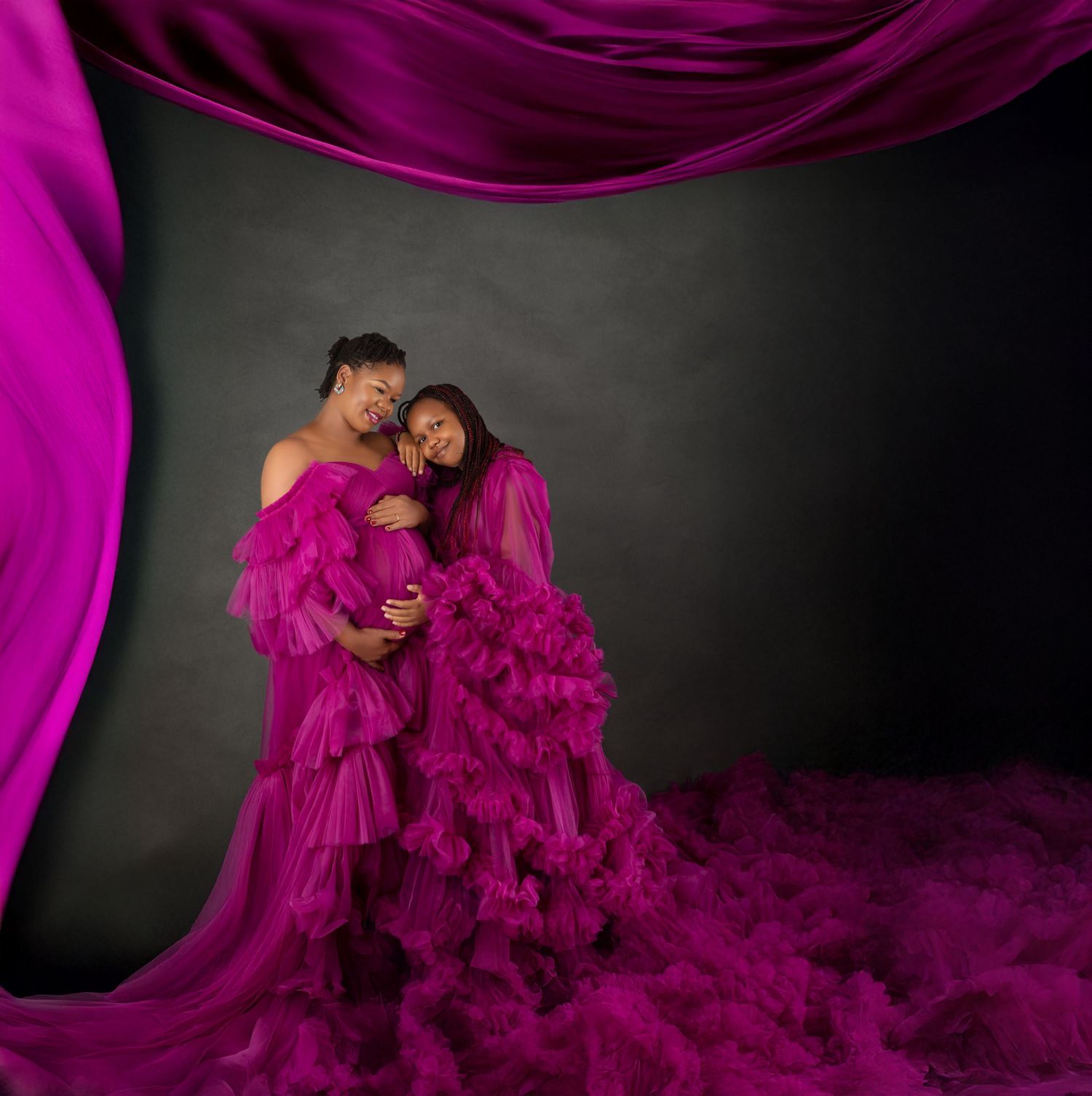Two women in flowing magenta tulle gowns pose together in a dramatic studio maternity portrait.