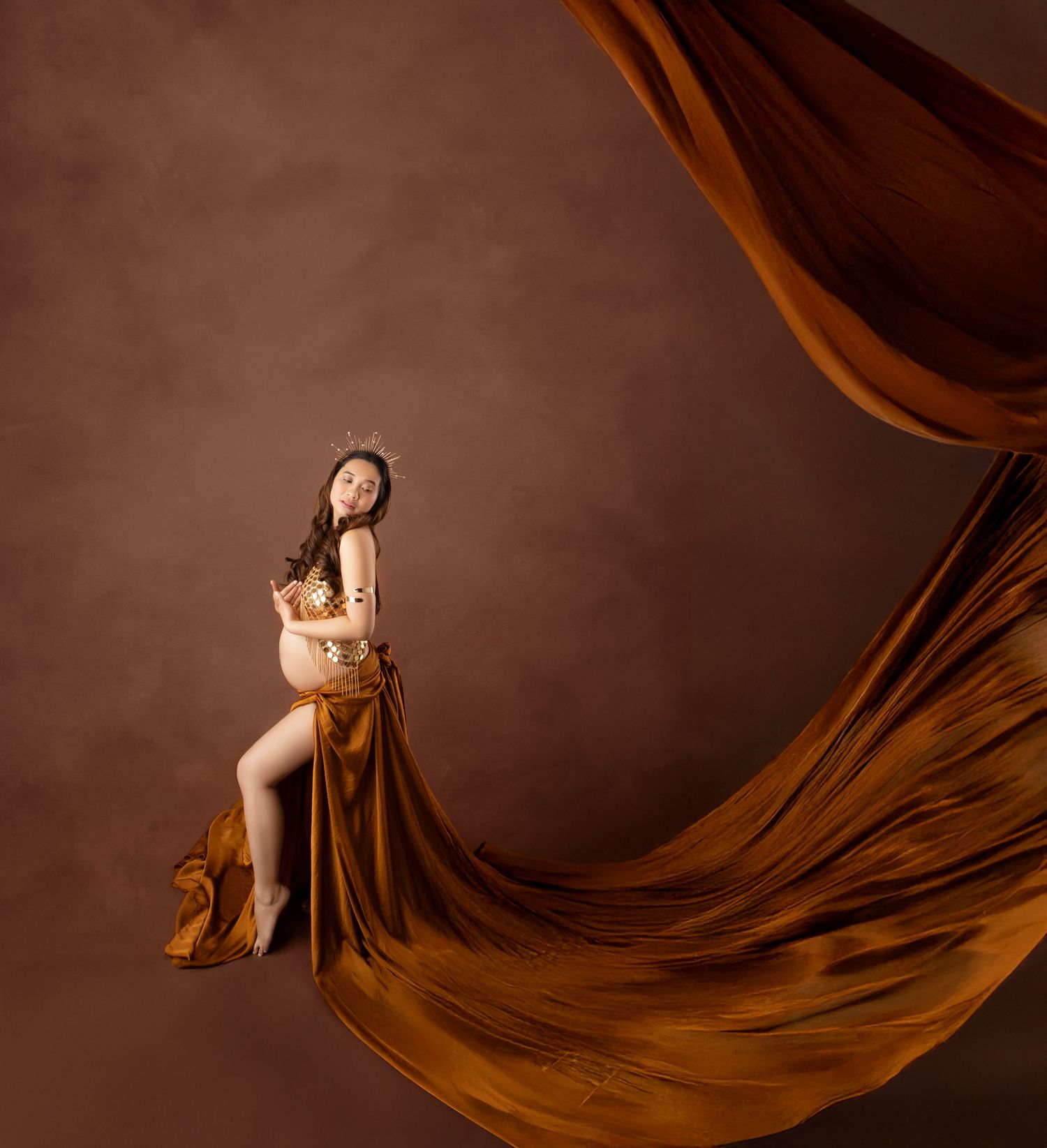 Pregnant woman in gold top and amber flowing fabric draped elegantly in warm-toned studio maternity photo.