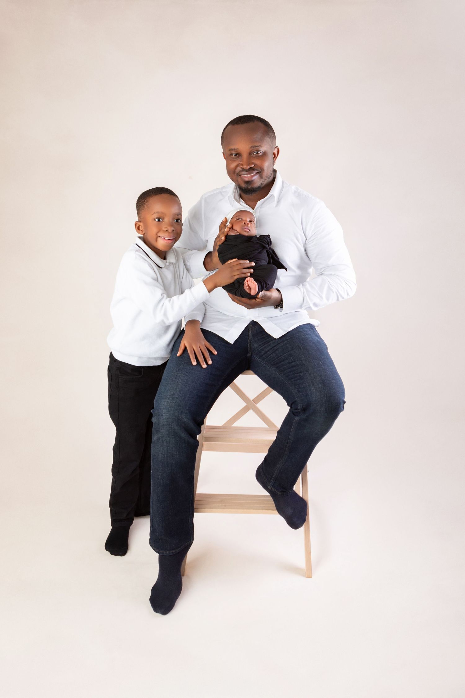 A father in a white shirt sits on a wooden stool holding a newborn while his young son stands beside him smiling.