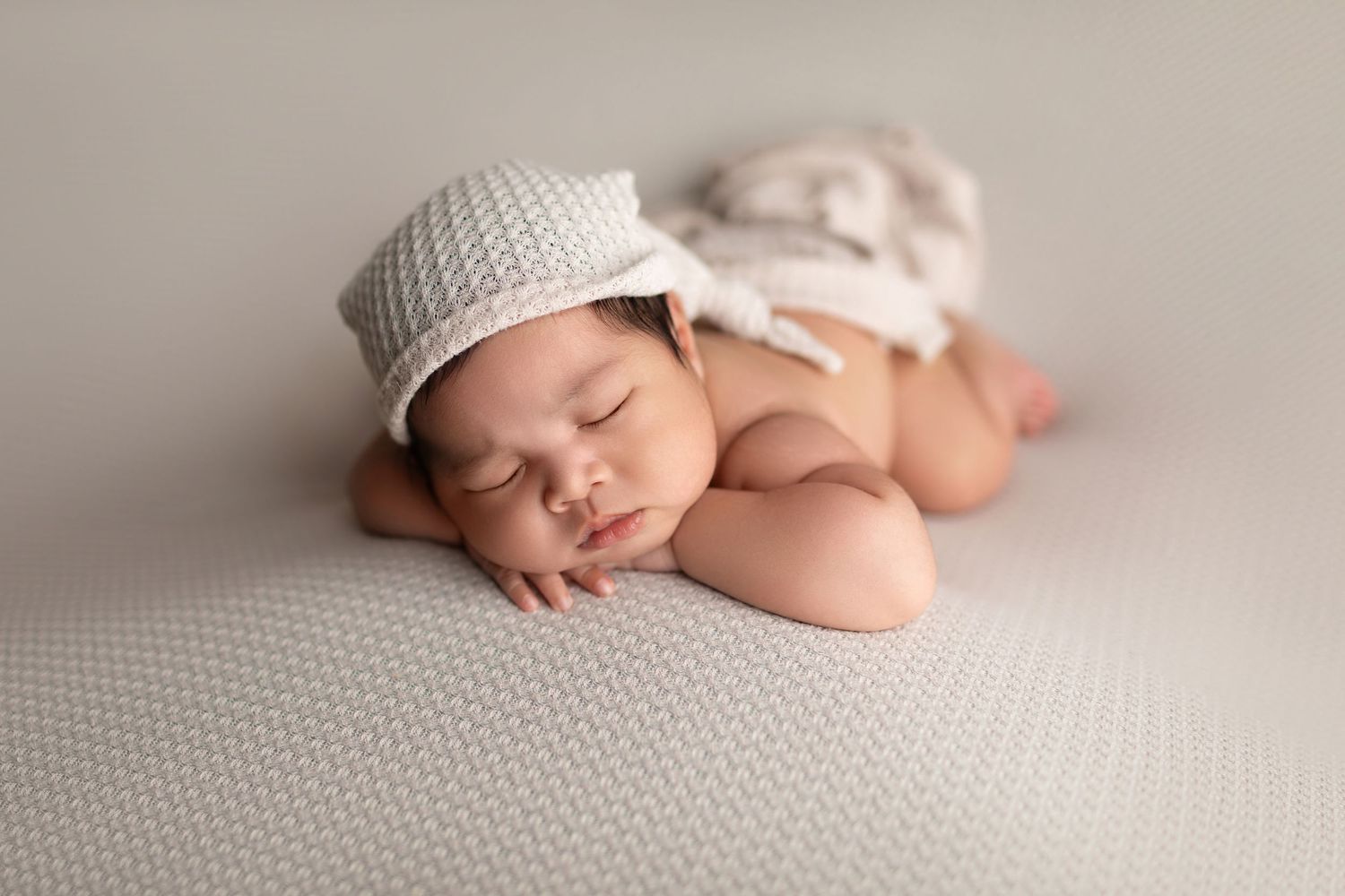 Sleeping newborn baby wearing a knit bonnet and diaper cover posed on a soft gray blanket.