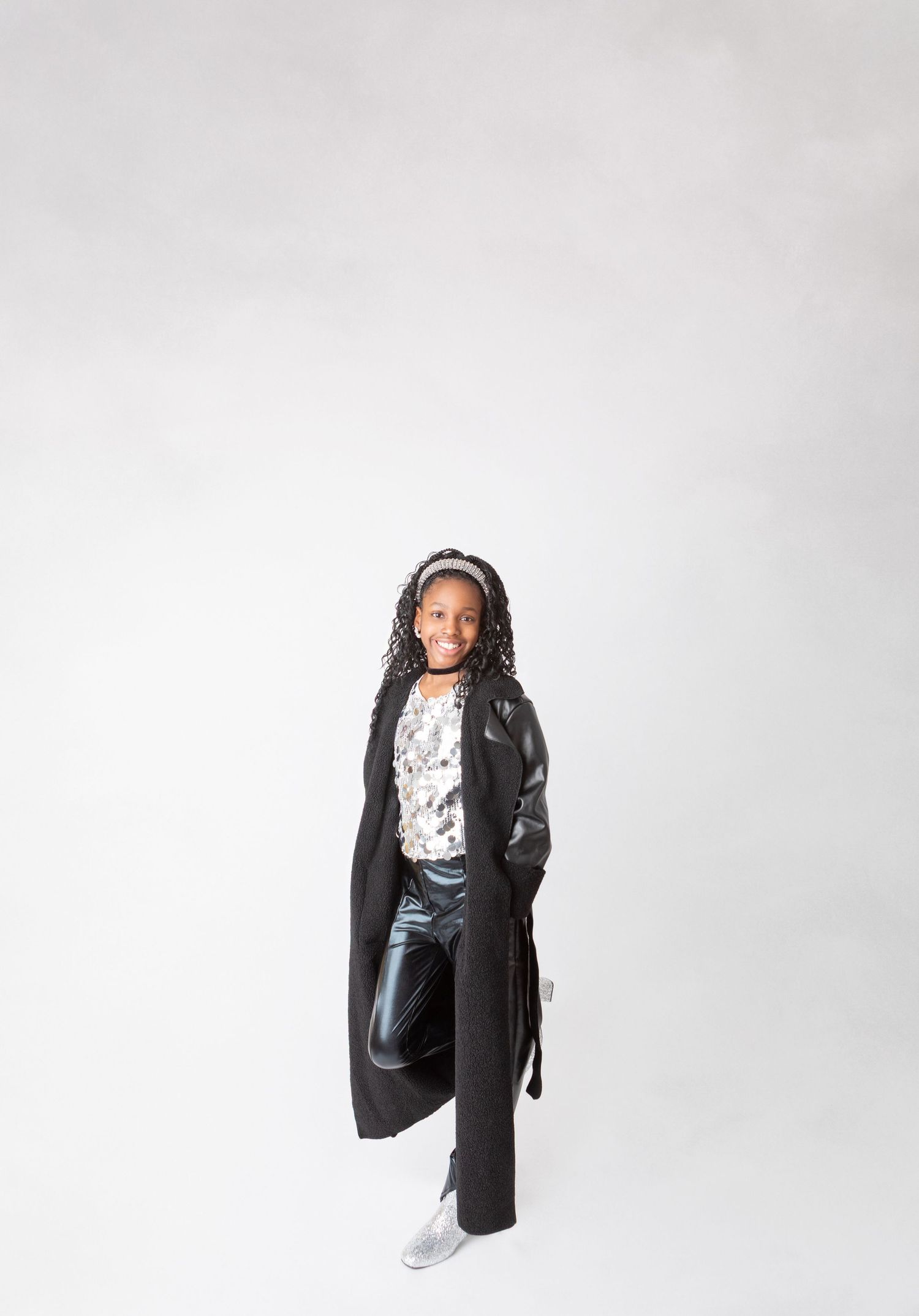 Young girl in black long coat, metallic pants, and silver shoes poses confidently against white background.
