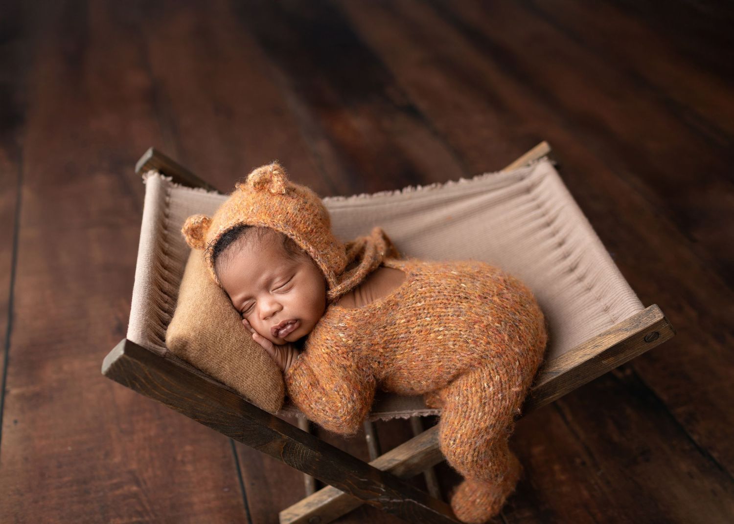 Newborn in brown teddy bear outfit sleeping in a tiny wooden crib.