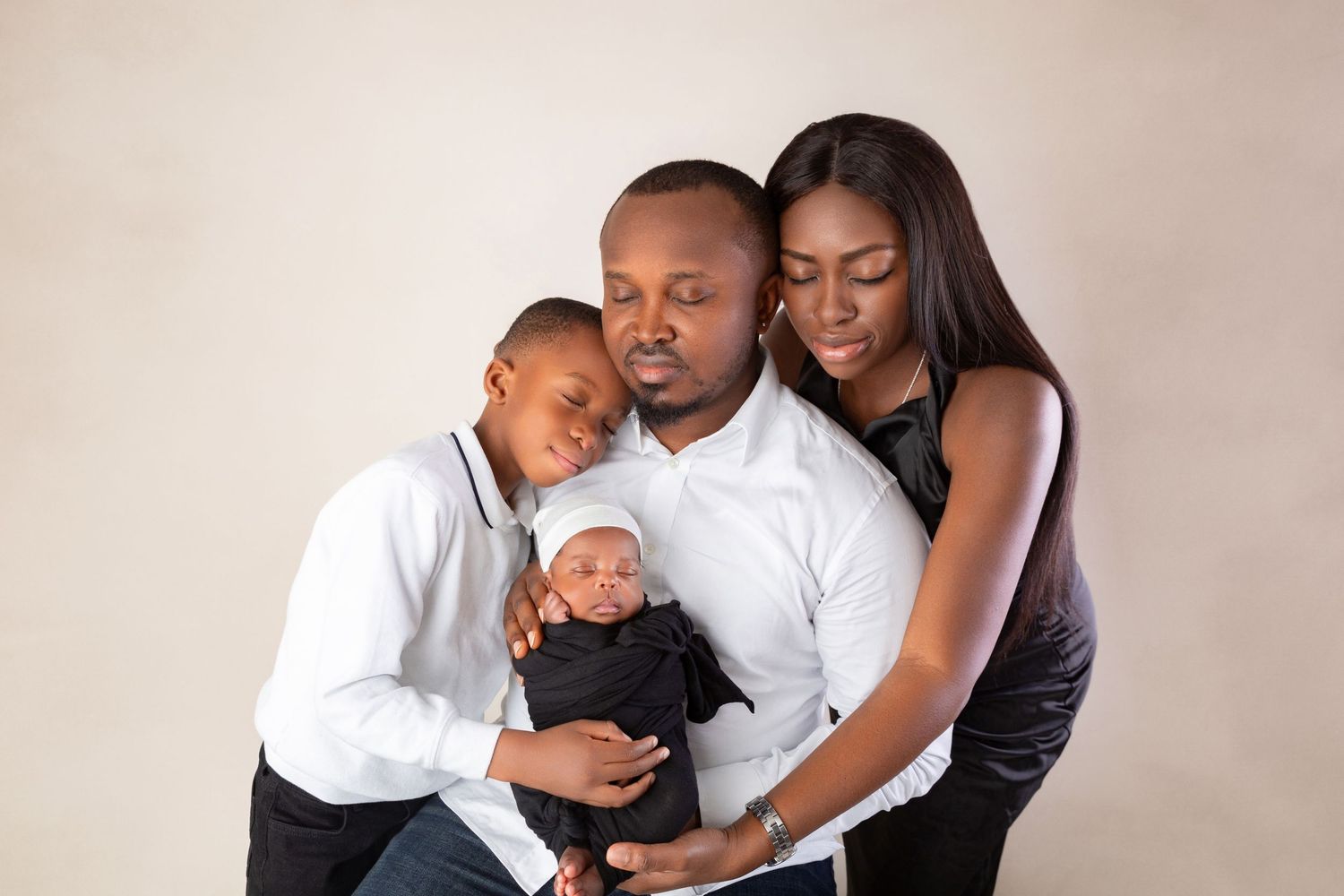 A family of four poses together in a studio, parents holding their newborn baby and young son in formal attire.
