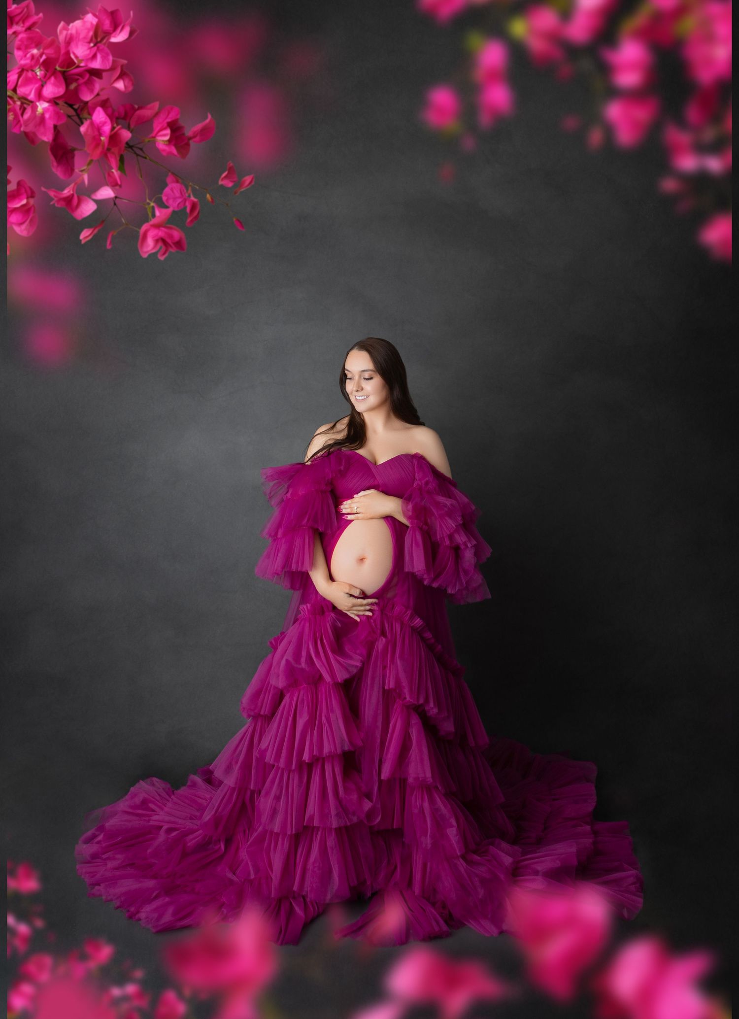 Pregnant woman in vibrant magenta ruffled tulle gown poses with pink flowers in dark studio setting.
