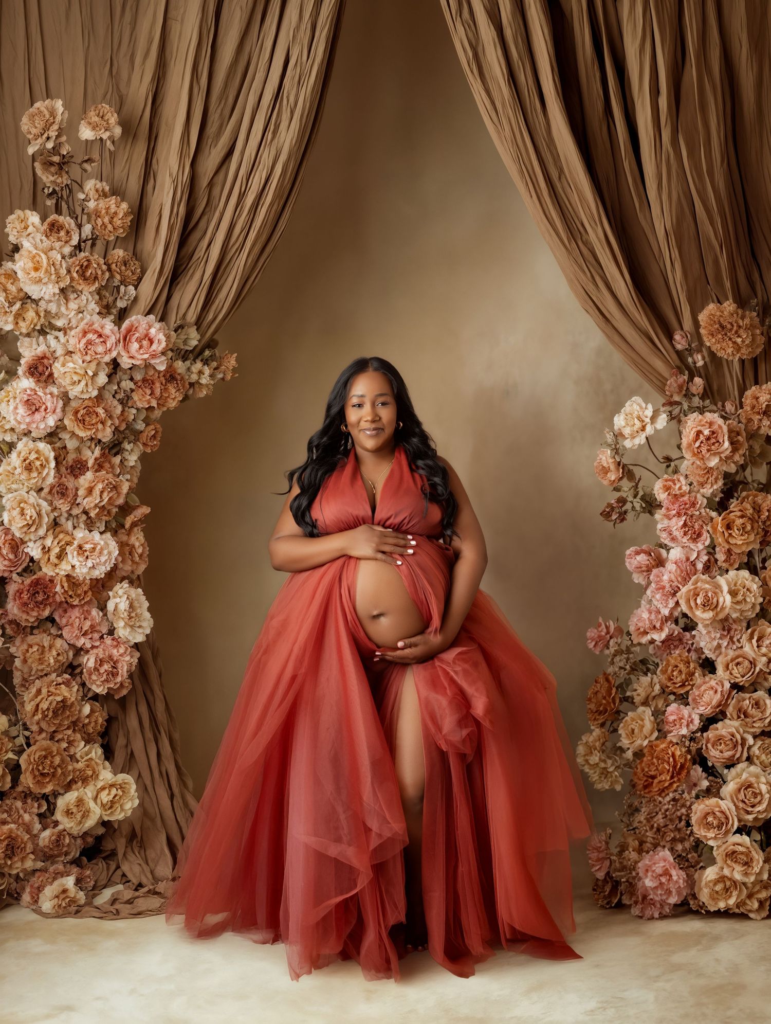 Pregnant woman in red tulle gown poses between floral columns and draped curtains in elegant maternity portrait.