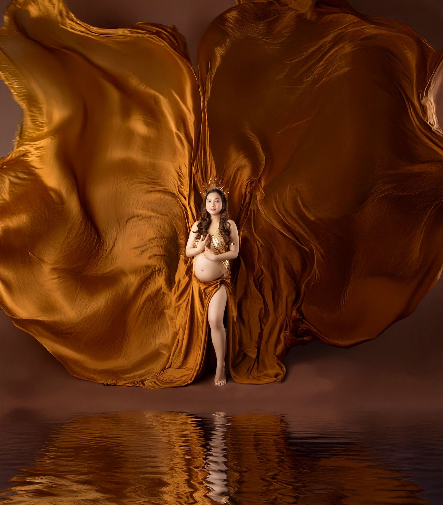 Pregnant woman in golden silk fabric flowing dramatically around her, reflected in water below, studio maternity photo.