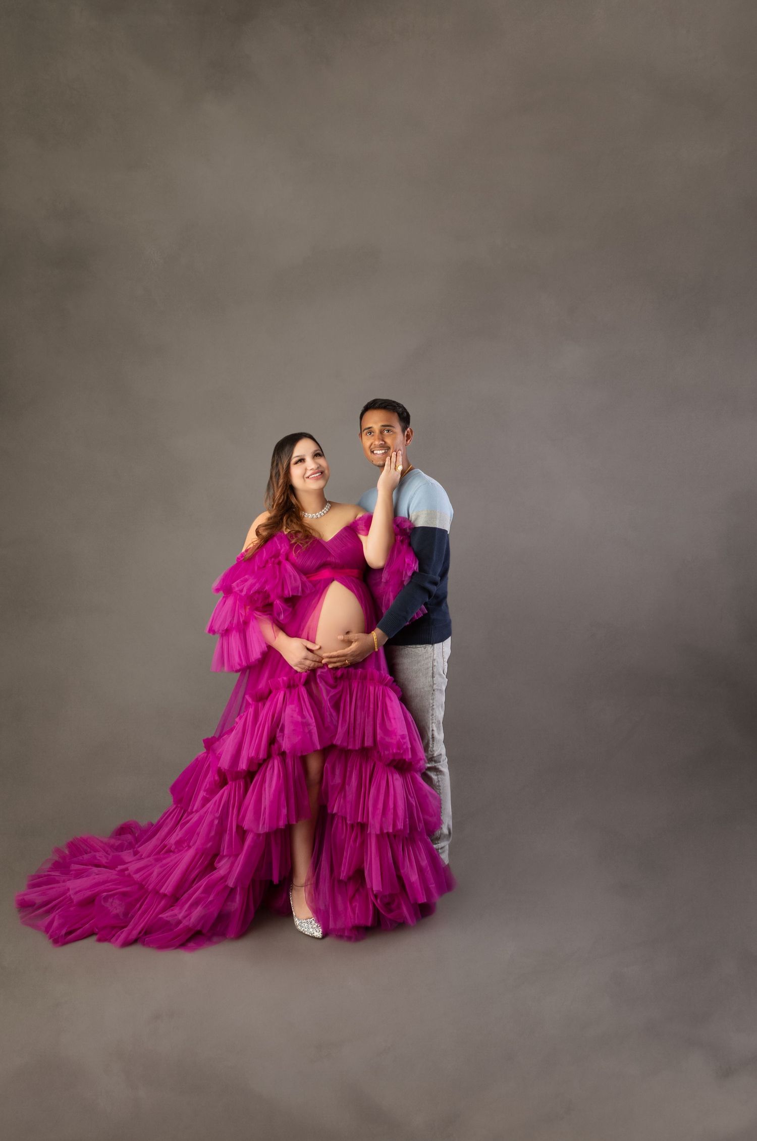 Smiling pregnant woman in fuchsia ruffled maternity gown posing with partner during studio photoshoot.