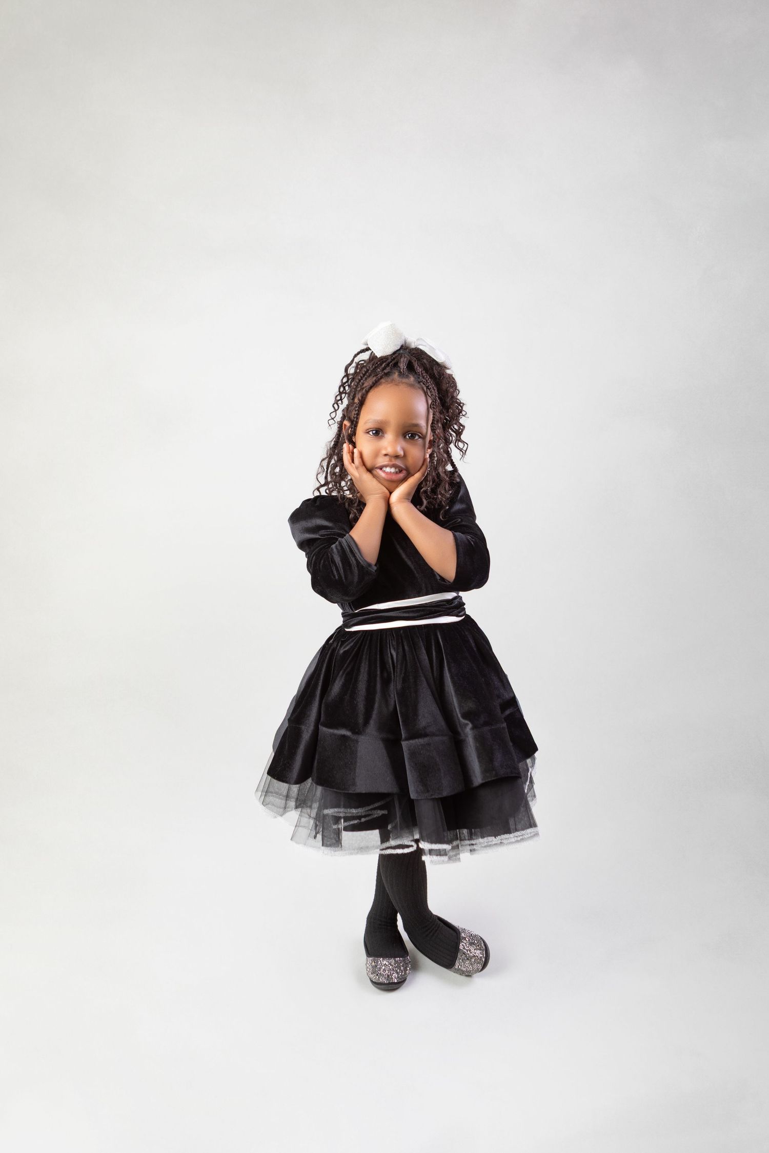 Adorable young Black girl in a black tiered dress with tulle, posing with hands on cheeks, smiling at camera.