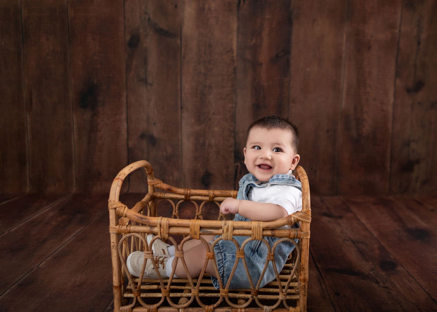 Happy baby boy sitting in a wicker bassinet against a rustic wood backdrop, smiling joyfully.