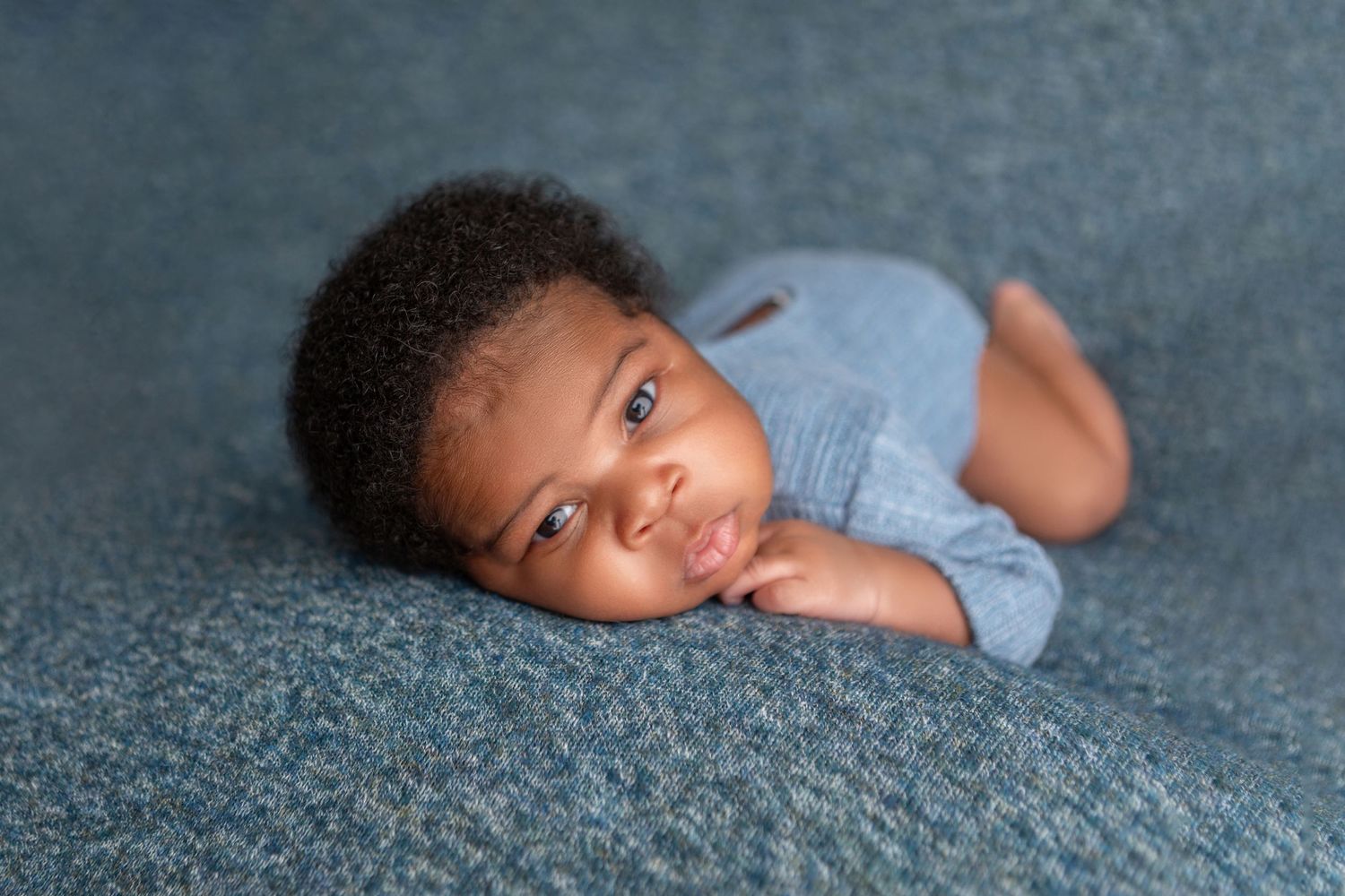 Newborn baby boy lying on a teal blanket wearing a blue romper, looking at the camera.