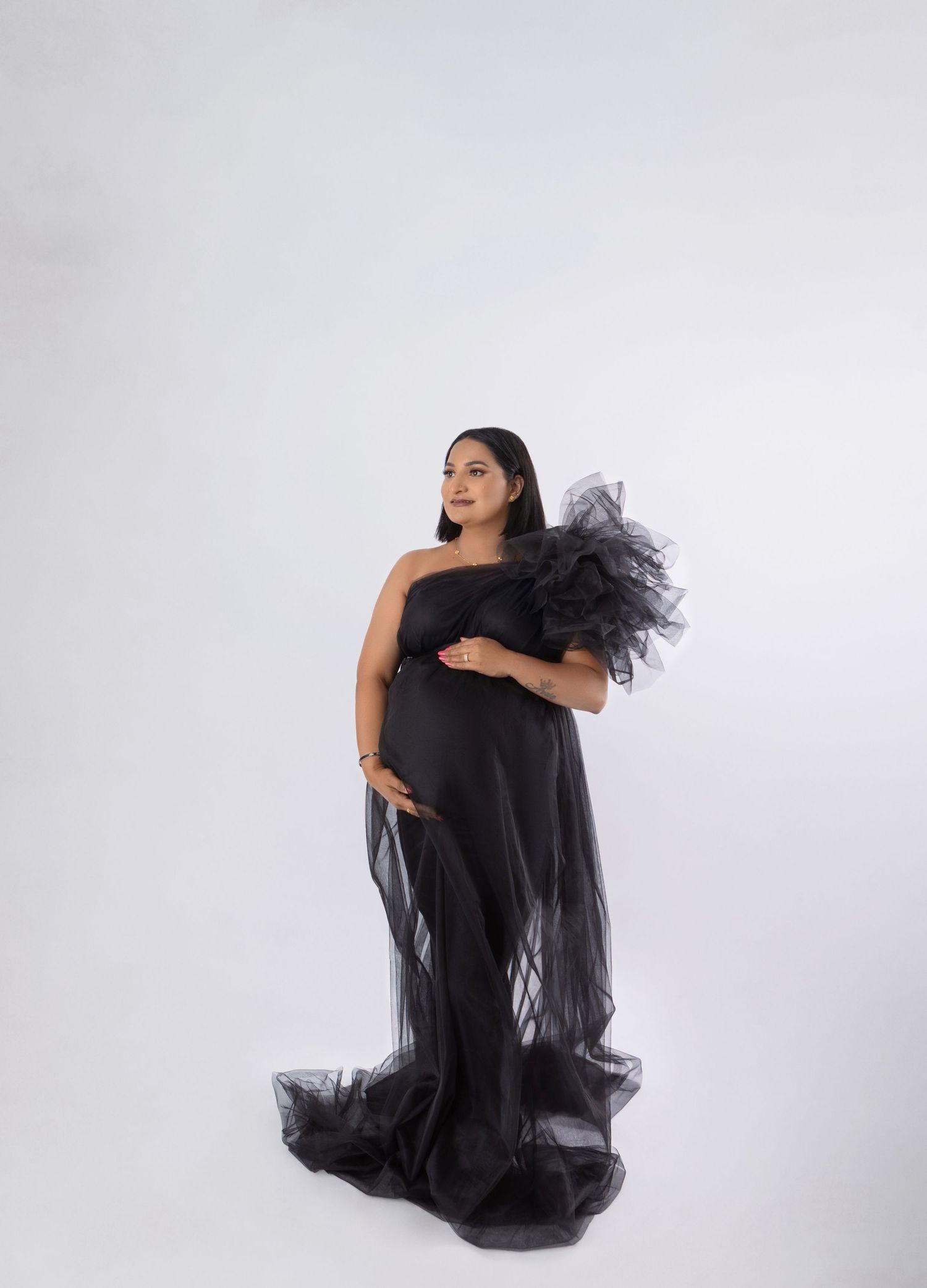Pregnant woman in elegant black one-shoulder tulle gown with ruffled detail, posing gracefully against white background.