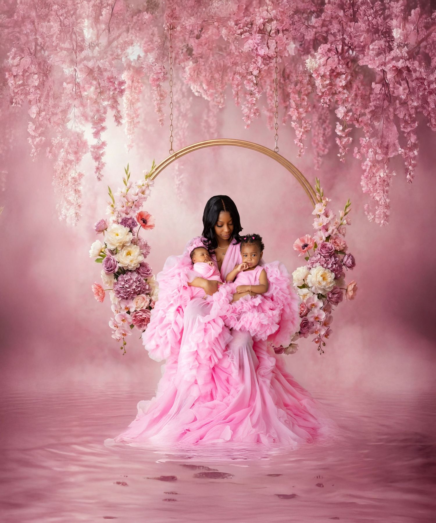 Mother in pink gown holds two babies under floral arch with hanging pink blossoms in dreamy studio portrait.