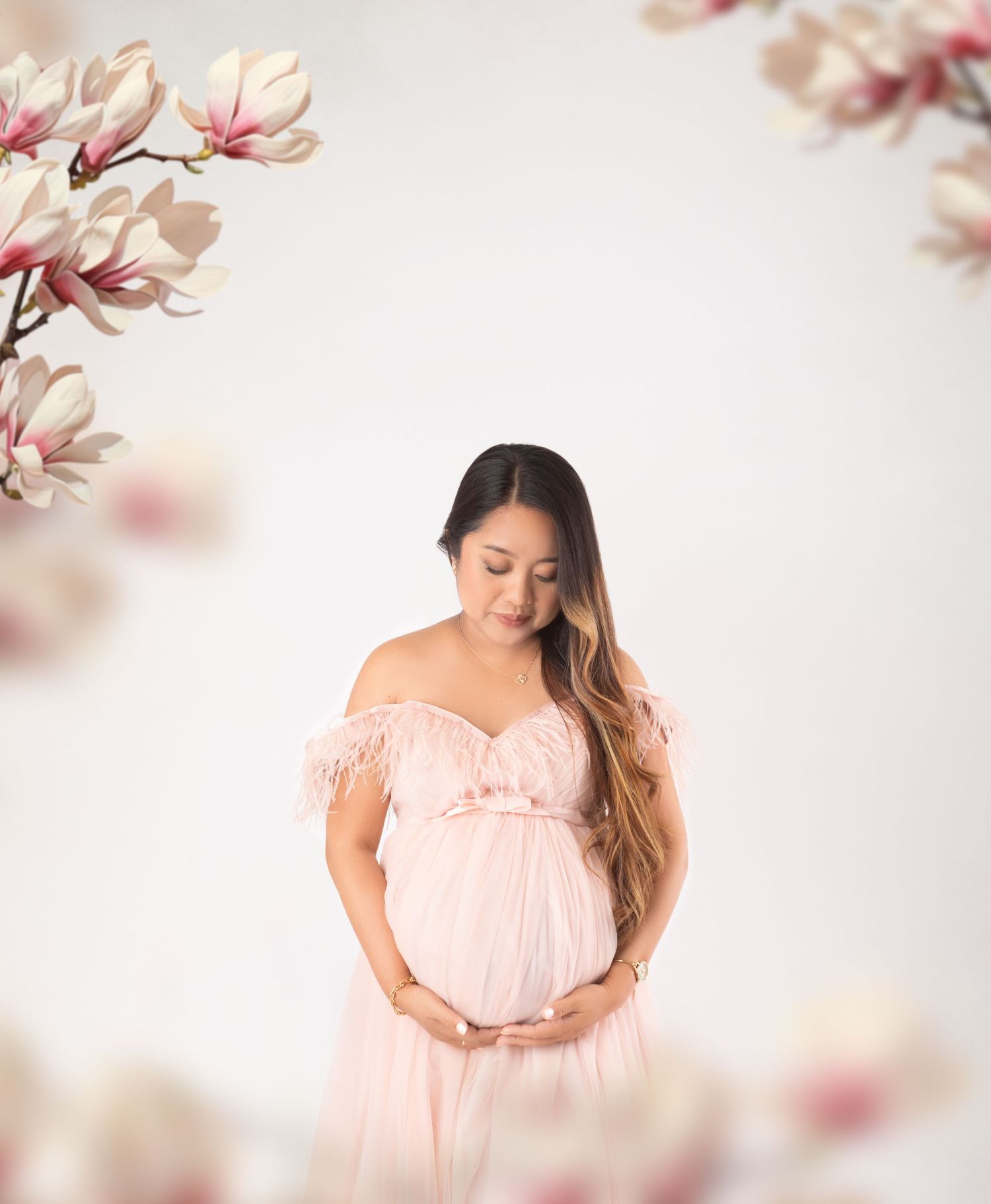 Pregnant woman in pink off-shoulder gown cradling baby bump, surrounded by blooming magnolia flowers on white background.