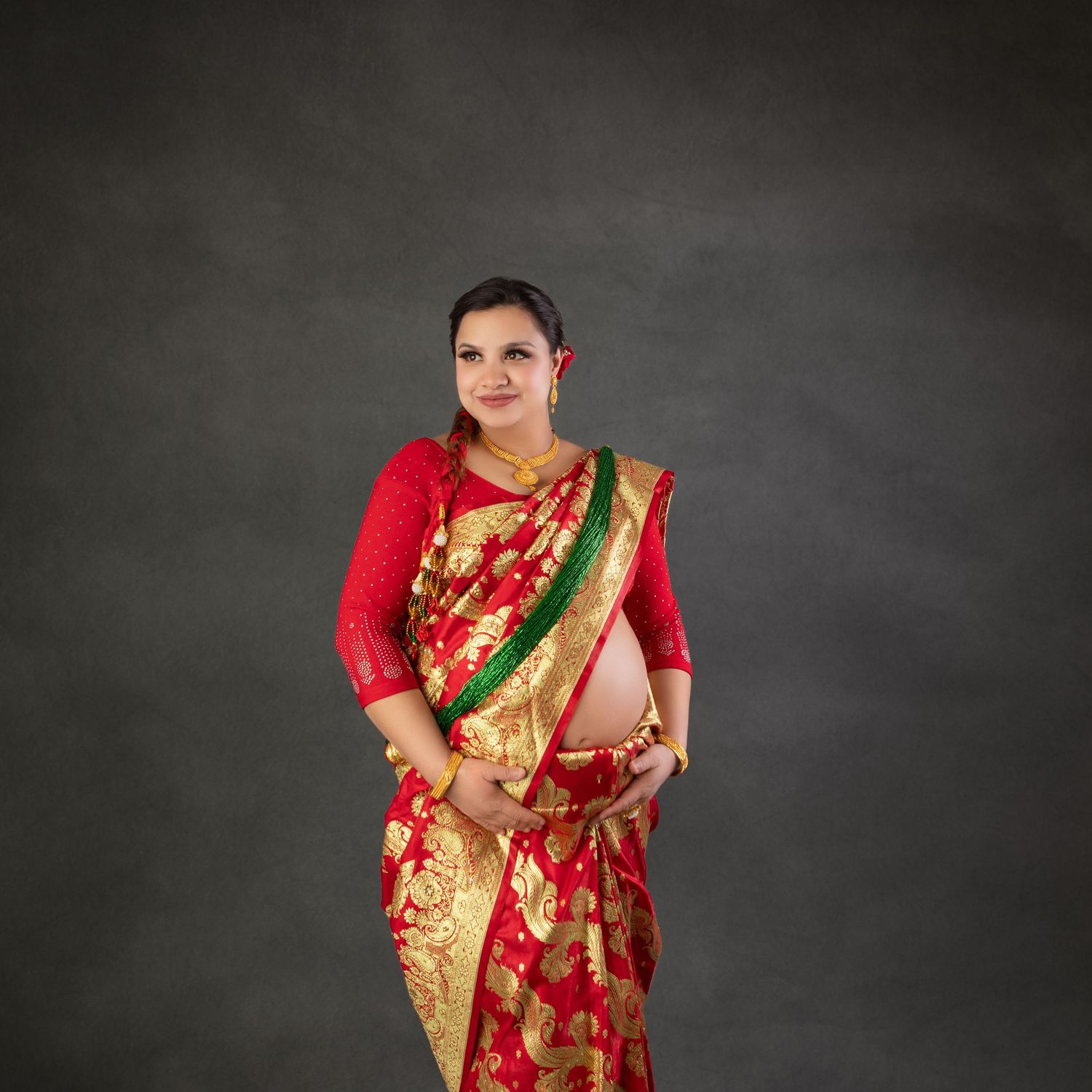 Pregnant woman in red and gold saree with green border, posing gracefully in a maternity photoshoot studio.