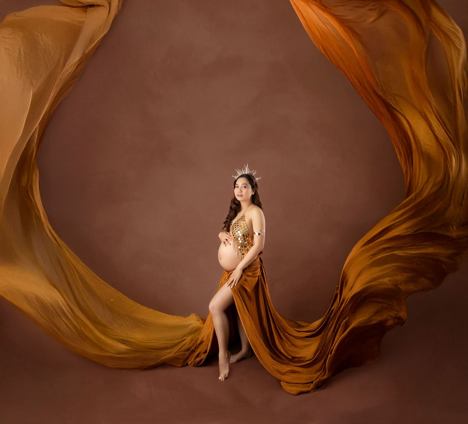 Pregnant woman in golden gown with crown poses gracefully against brown backdrop with flowing fabric wings.
