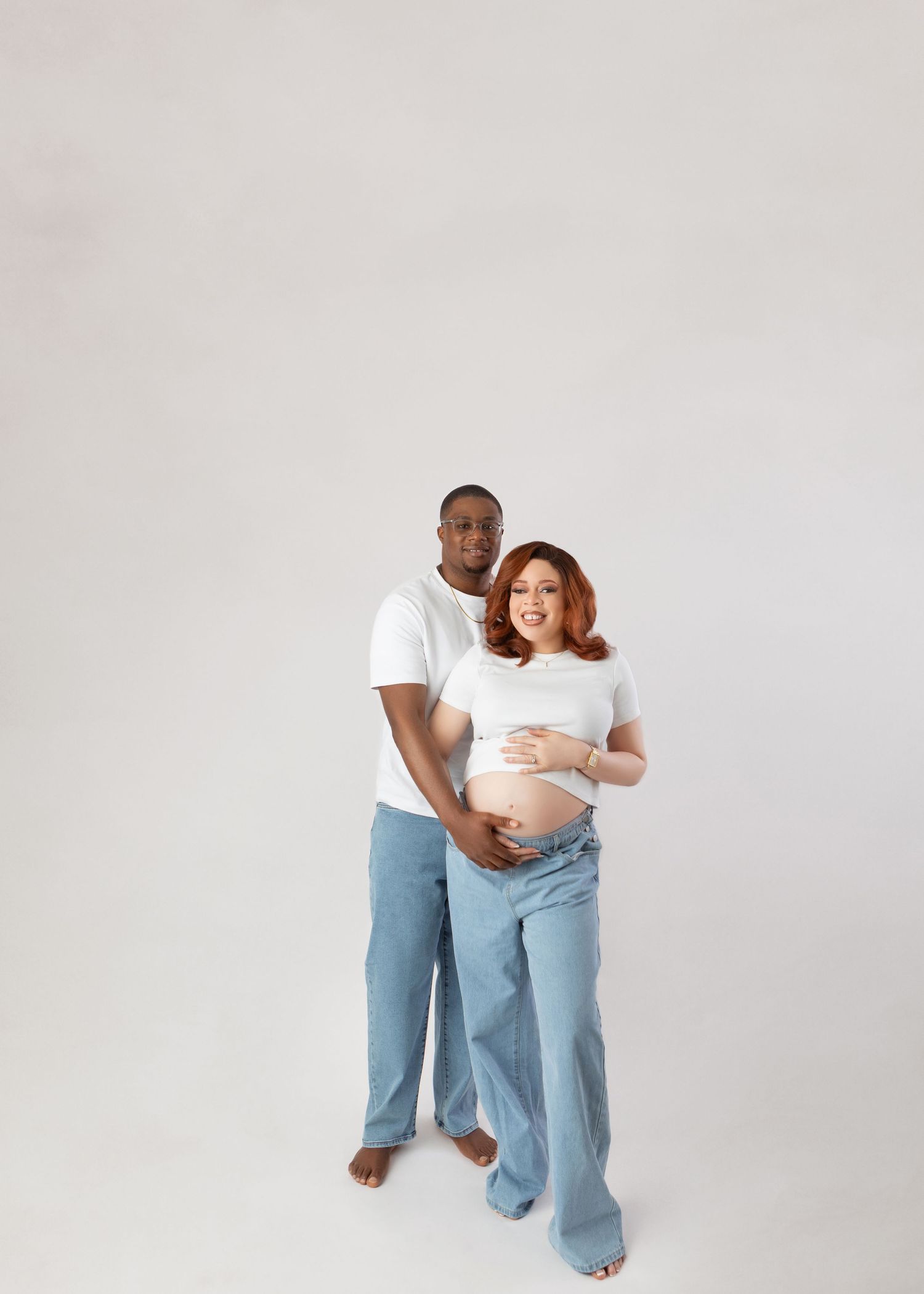 Maternity couple portrait in white tops and jeans, man embracing pregnant woman from behind on white backdrop.