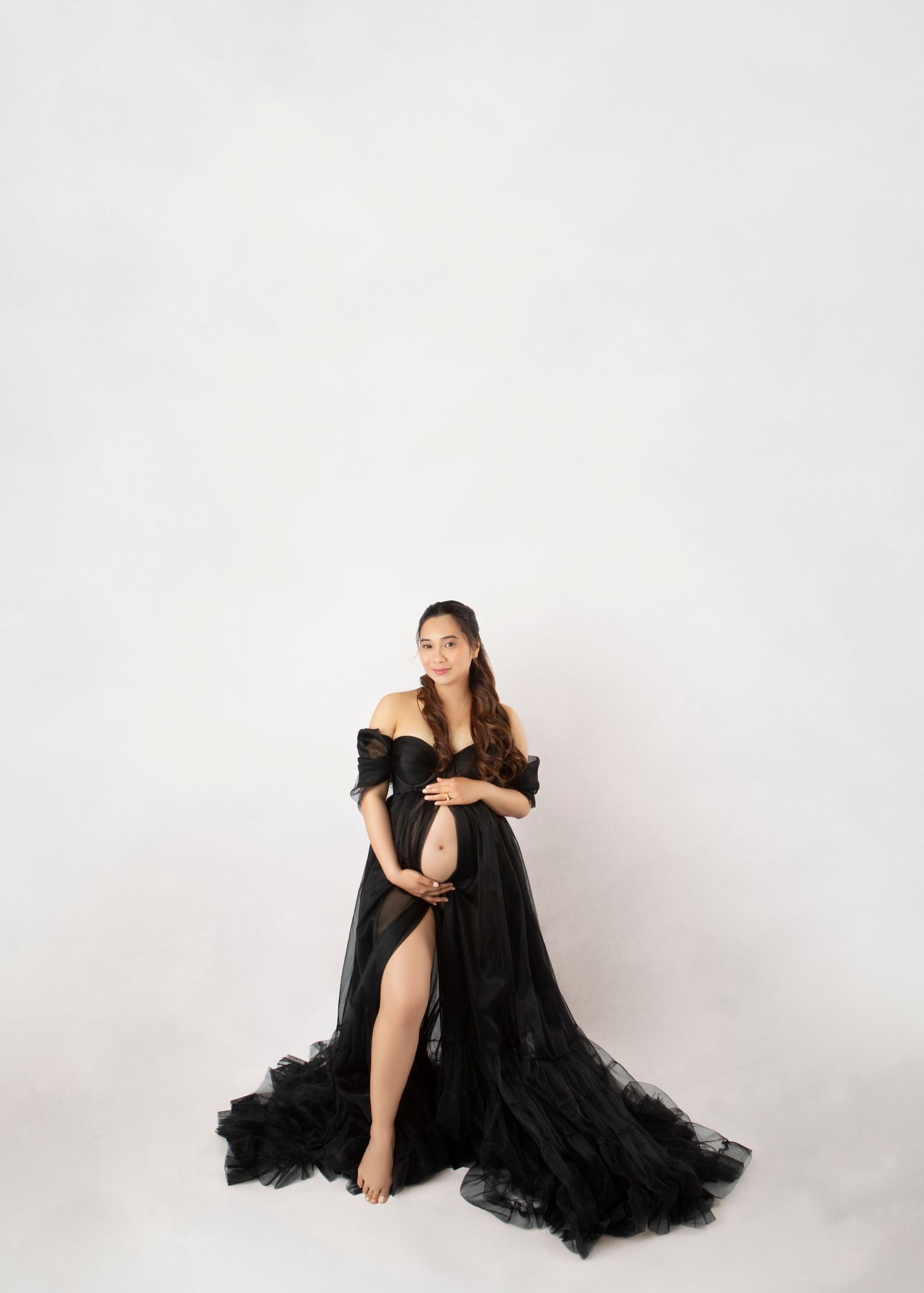 A pregnant woman in a sheer black off-shoulder tulle maternity gown poses against a white backdrop.