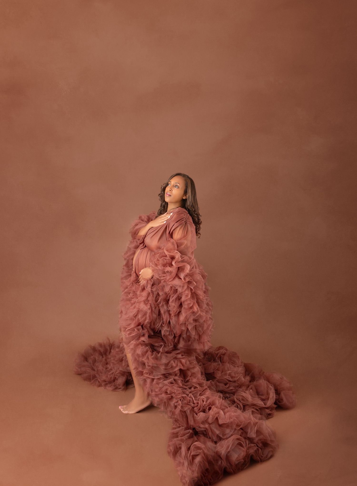 Pregnant woman in flowing dusty rose ruffled gown gazes upward in a warm brown studio maternity photo.