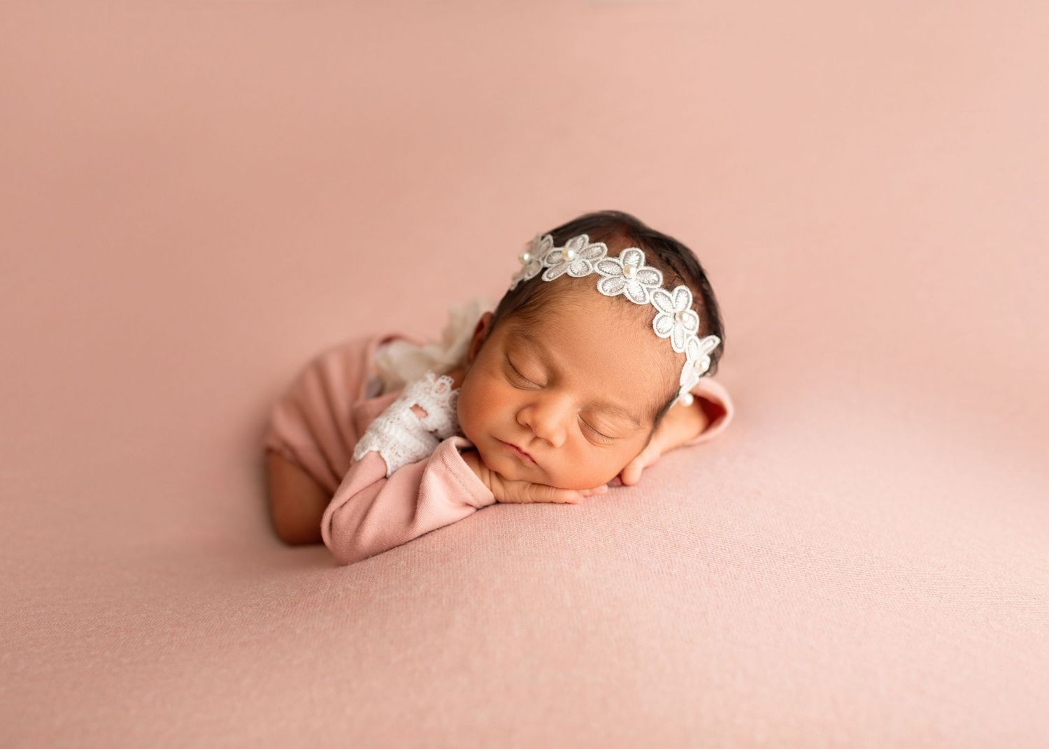 Sleeping newborn baby girl in pink outfit with white floral headband resting on a soft pink backdrop.