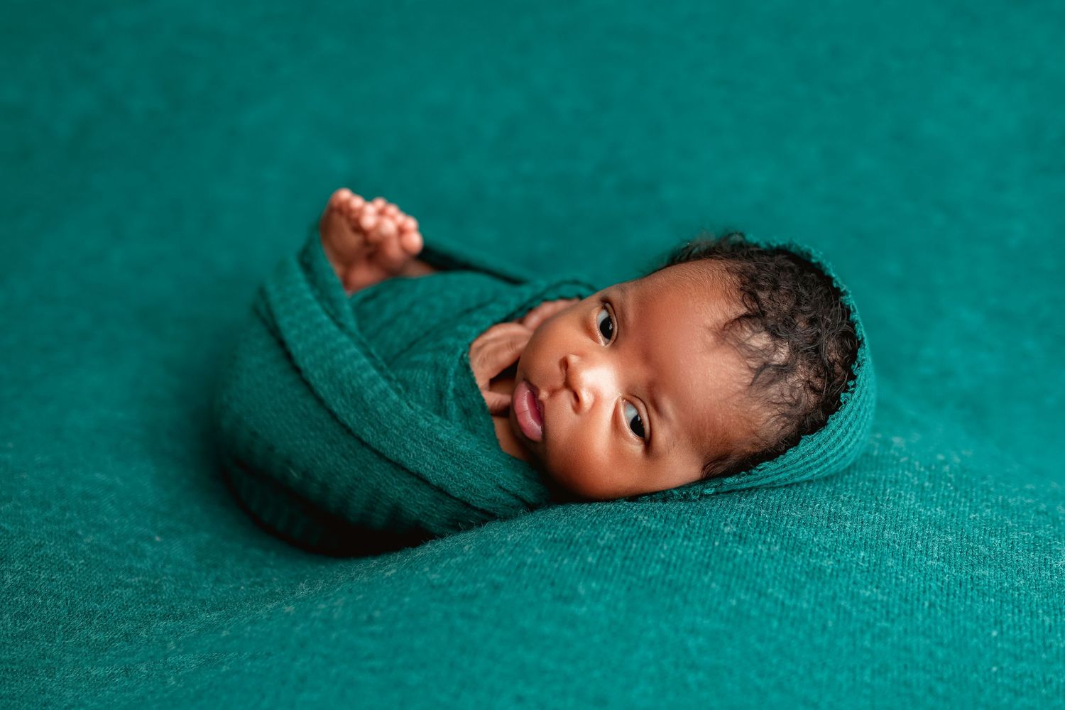 Alert newborn wrapped in teal fabric sticking out tongue on a teal textured backdrop.