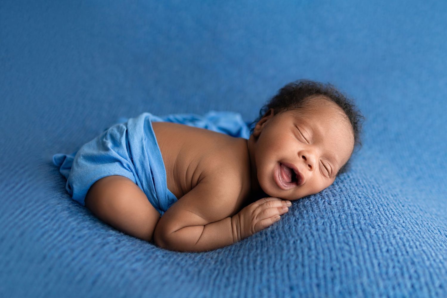 Smiling newborn baby wrapped in blue on a blue knit blanket.