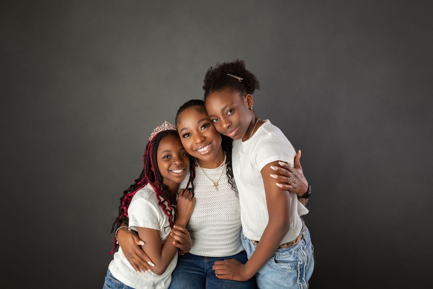 Three smiling Black sisters hugging closely in matching white tops and jeans against gray studio backdrop.