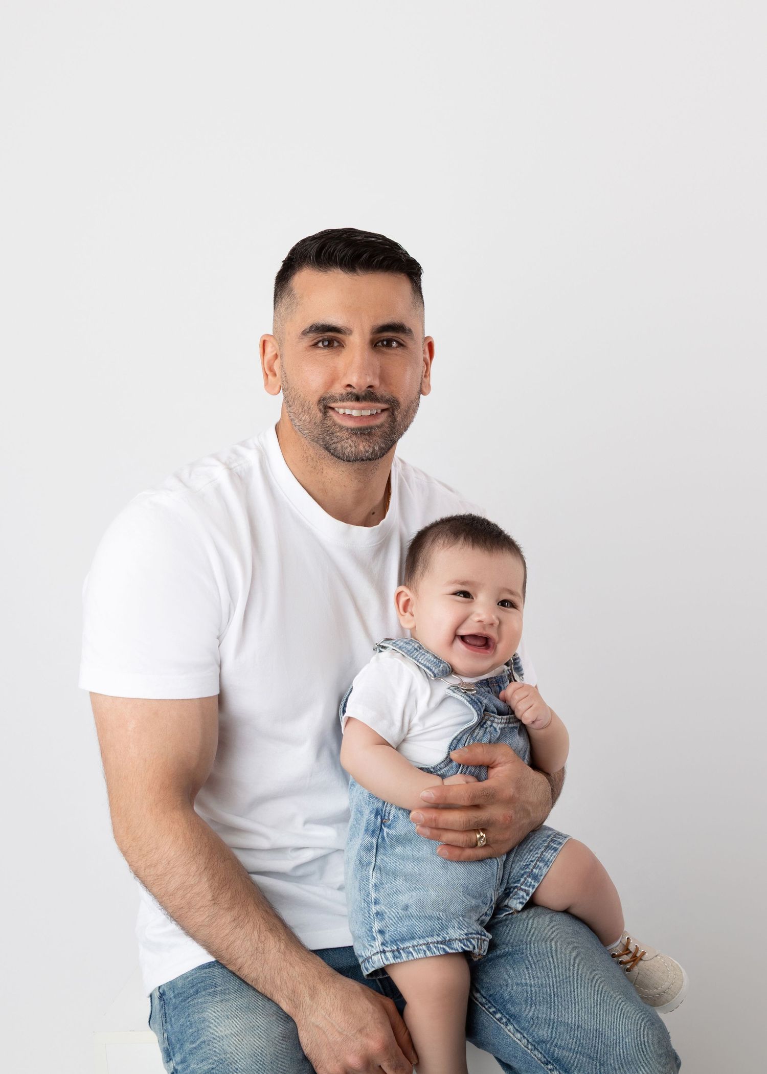 Smiling father holding laughing baby in denim overalls against white background.