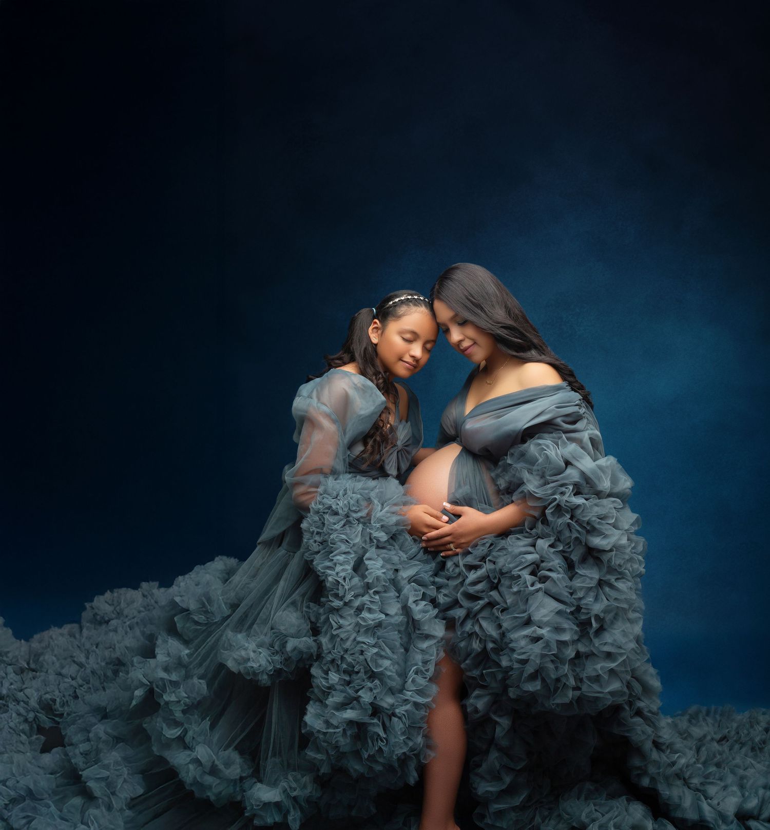 Pregnant woman and young girl in matching blue ruffle gowns pose together in a studio maternity photo shoot.